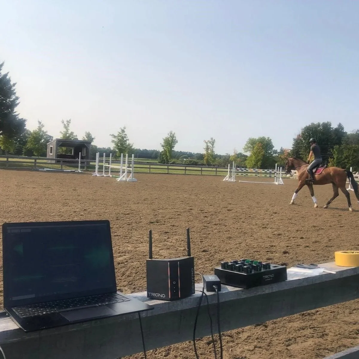 An outdoor riding arena with a person riding a horse, several jumps set up, and a laptop and Delsys Trigno surface electromyography system and sensors on a table in the foreground.