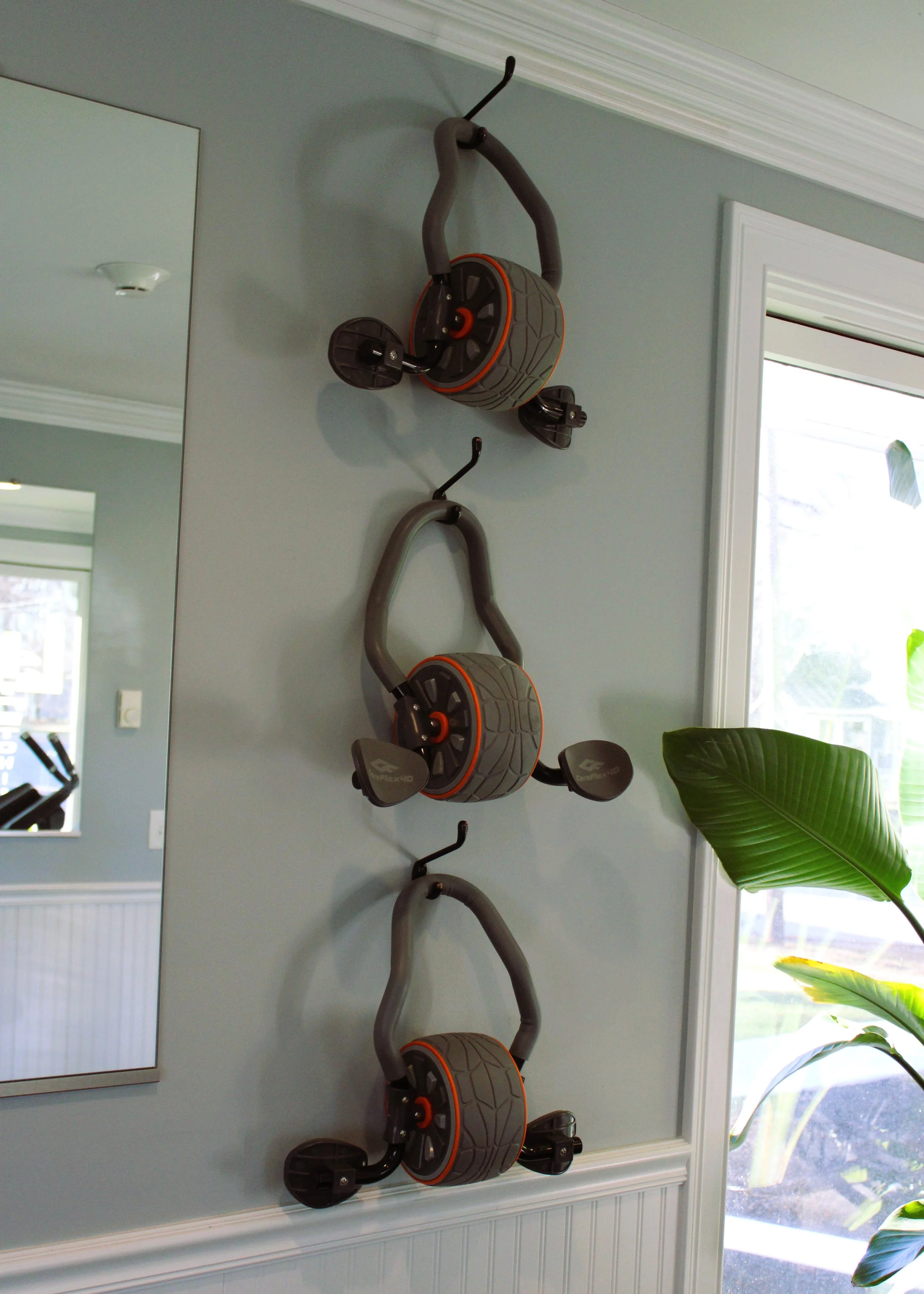Three gray and orange kids' scooters mounted on a light blue wall near a window and a mirror.