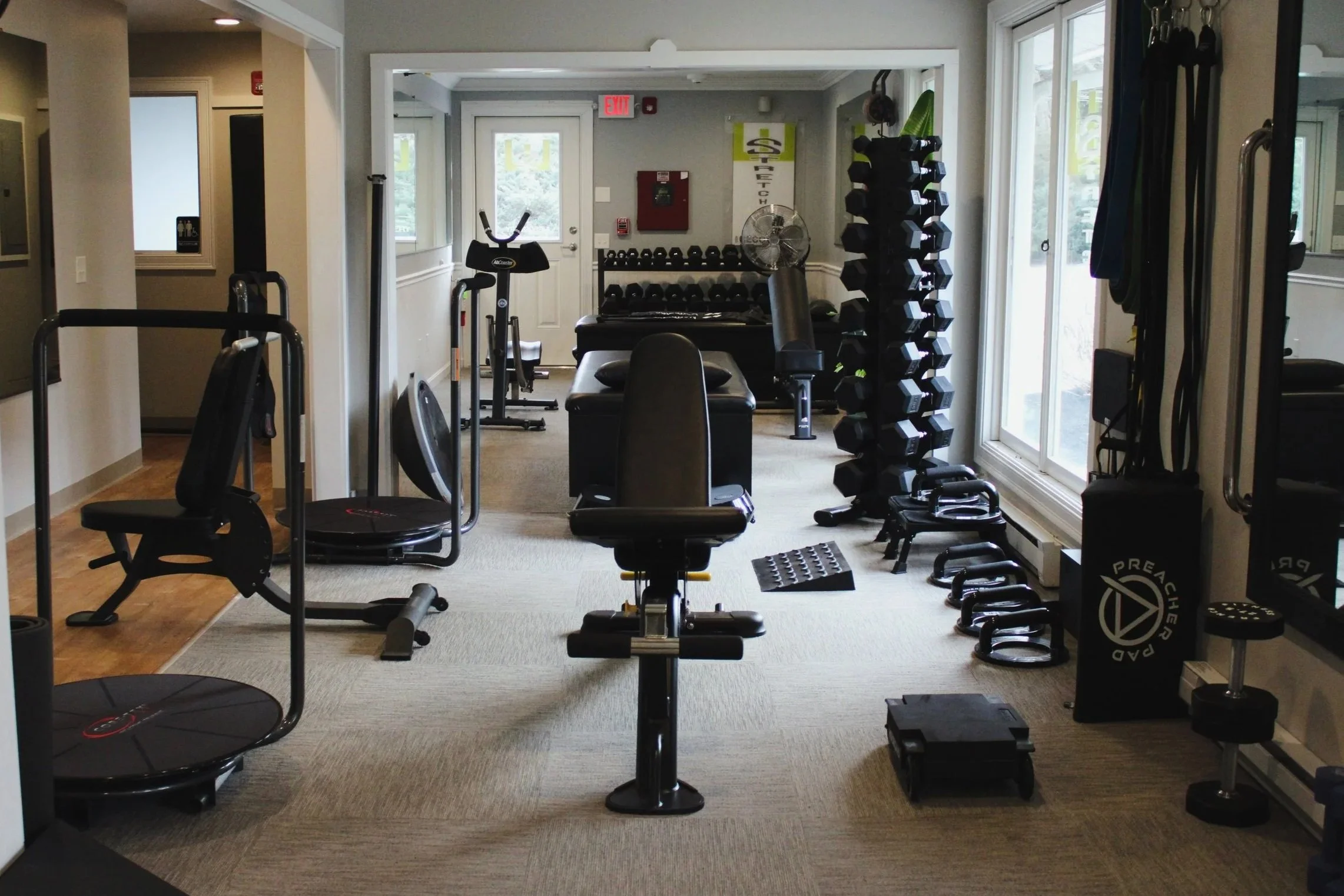 Indoor gym room with workout equipment including dumbbells, medicine balls, exercise bikes, a weight bench, kettlebells, and resistance bands, with windows and an exit door in the background.
