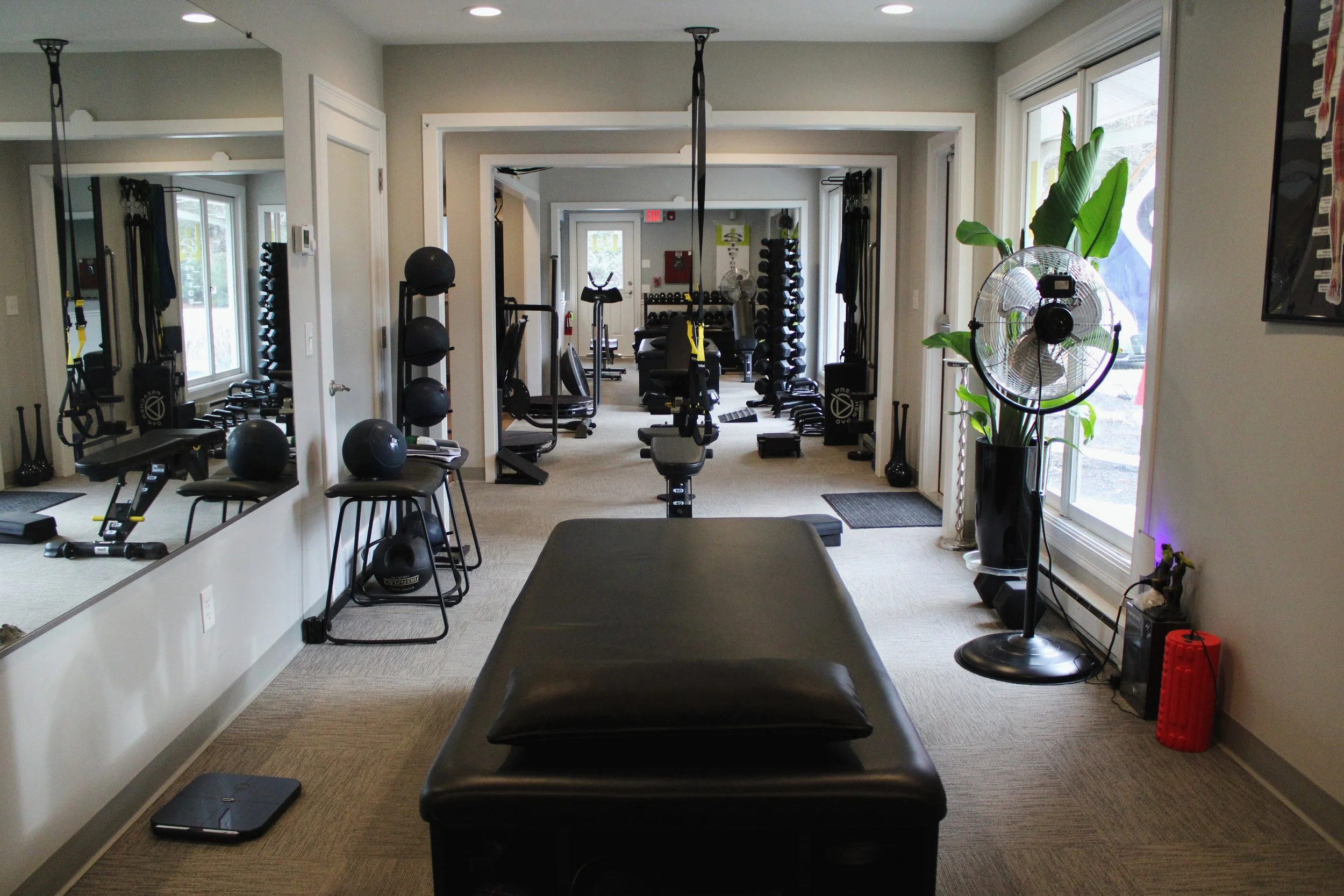 A therapy or consultation room with a padded black sofa, four black chairs, large wall mirror, two wall-mounted televisions displaying colorful logos or icons, and beige carpeting.