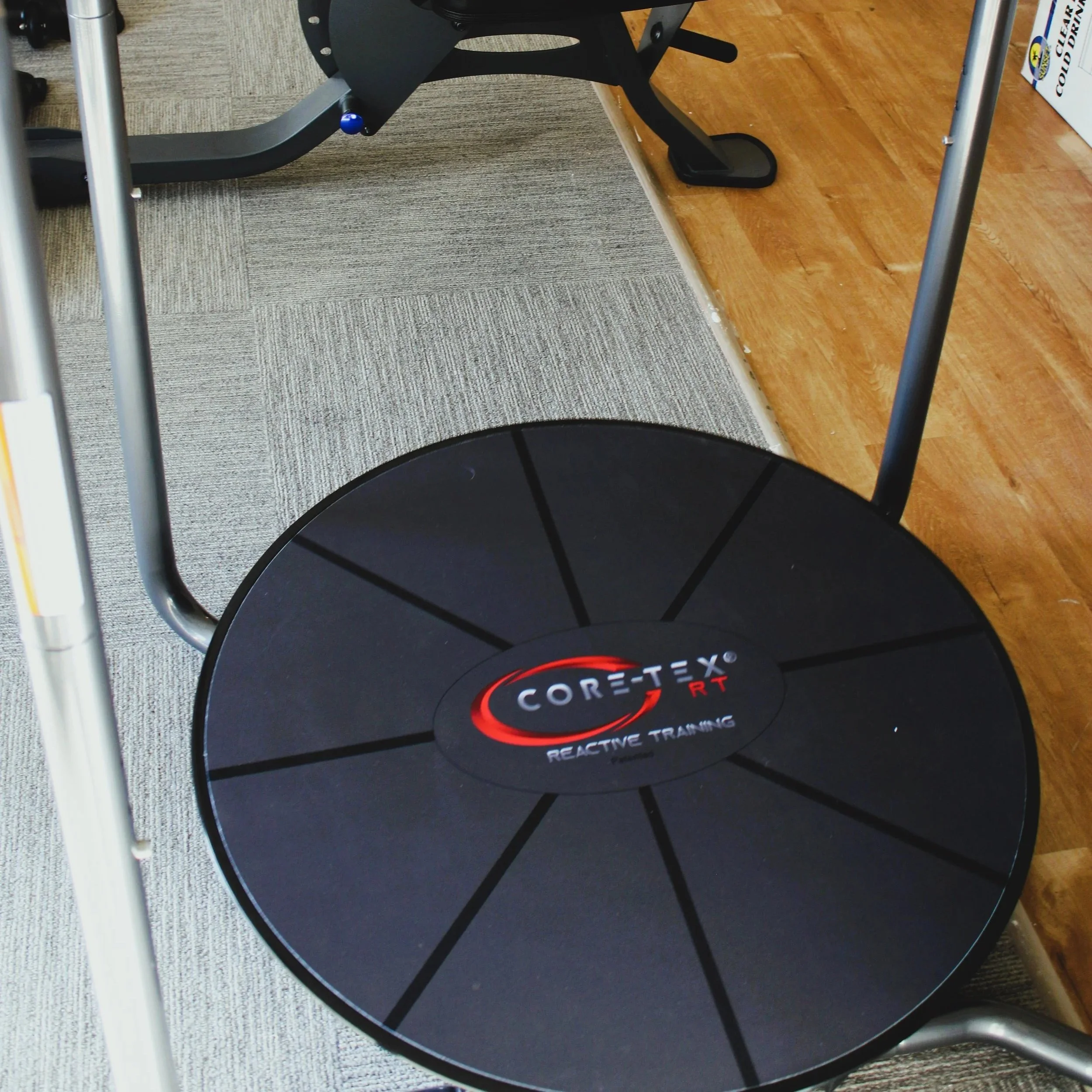 A black Core-Tex RT reactive training balance and stability platform with a circular shape and segmented design, placed on carpeted flooring near wooden floor in a gym or training space.