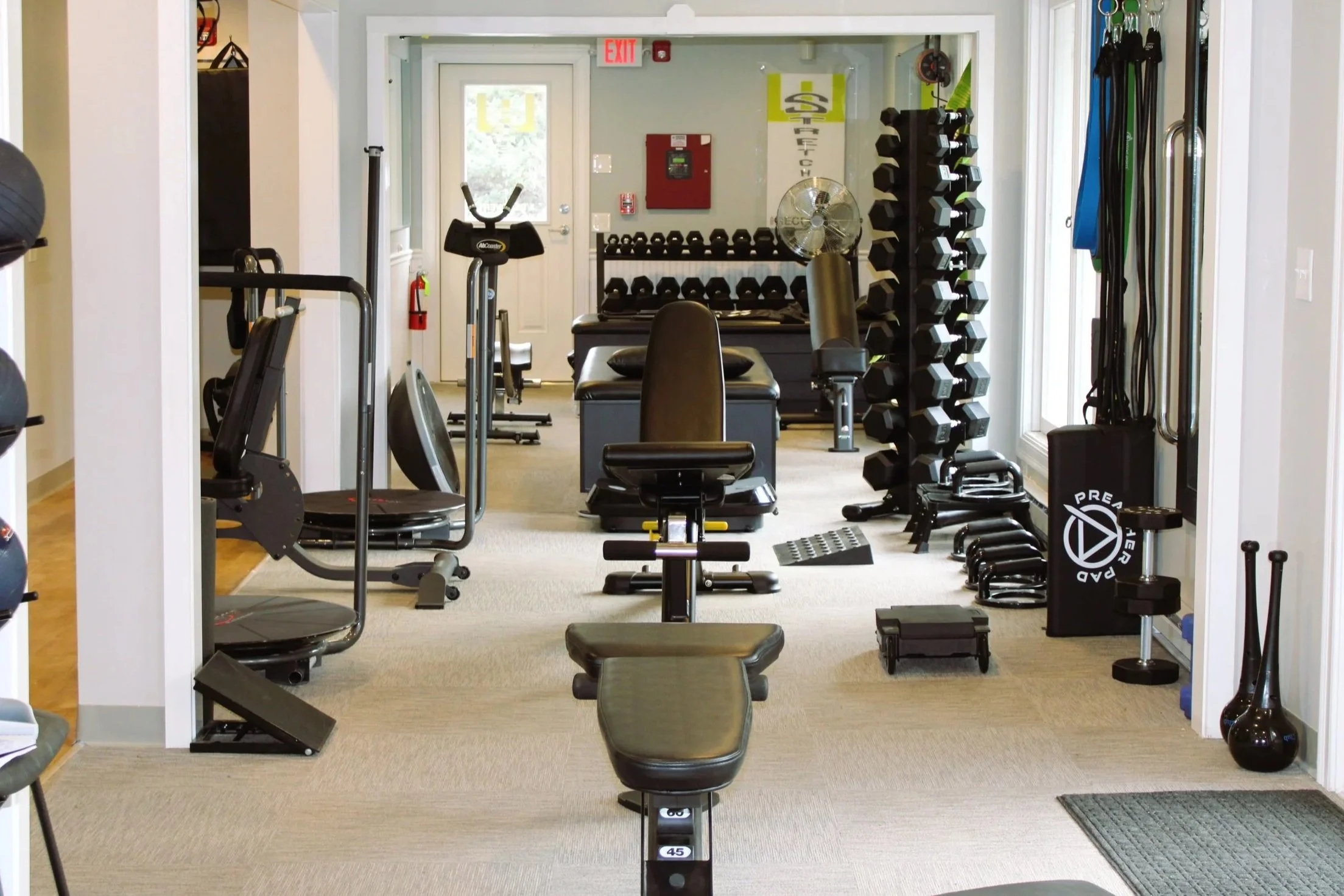 Empty gym with various workout equipment including a weight bench, dumbbells, kettlebells, and cardio machines, arranged neatly in a bright room with large windows.