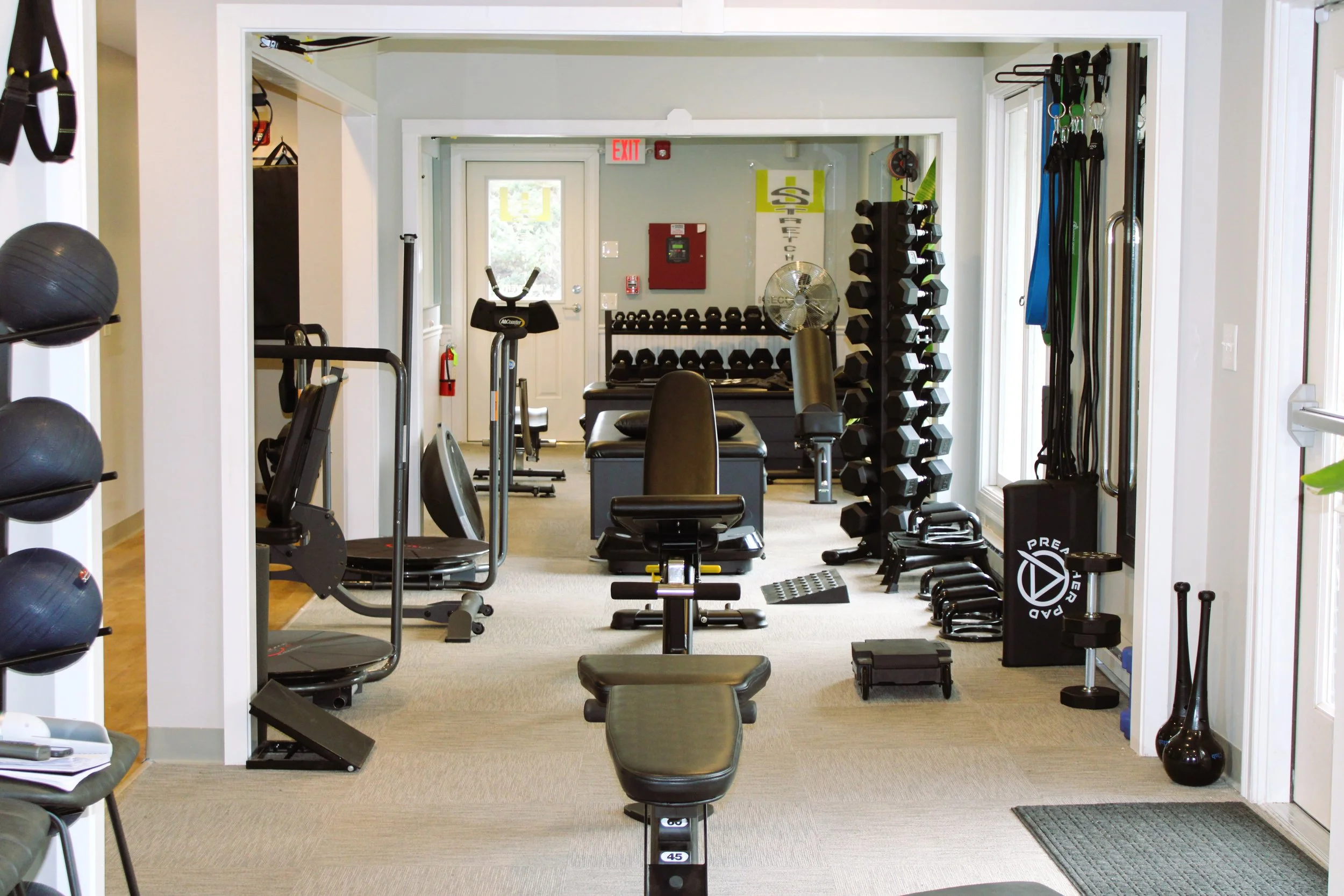 A small gym with various exercise equipment, including dumbbells, medicine balls, resistance bands, and a weight bench, organized in a well-lit room with windows on the right side.