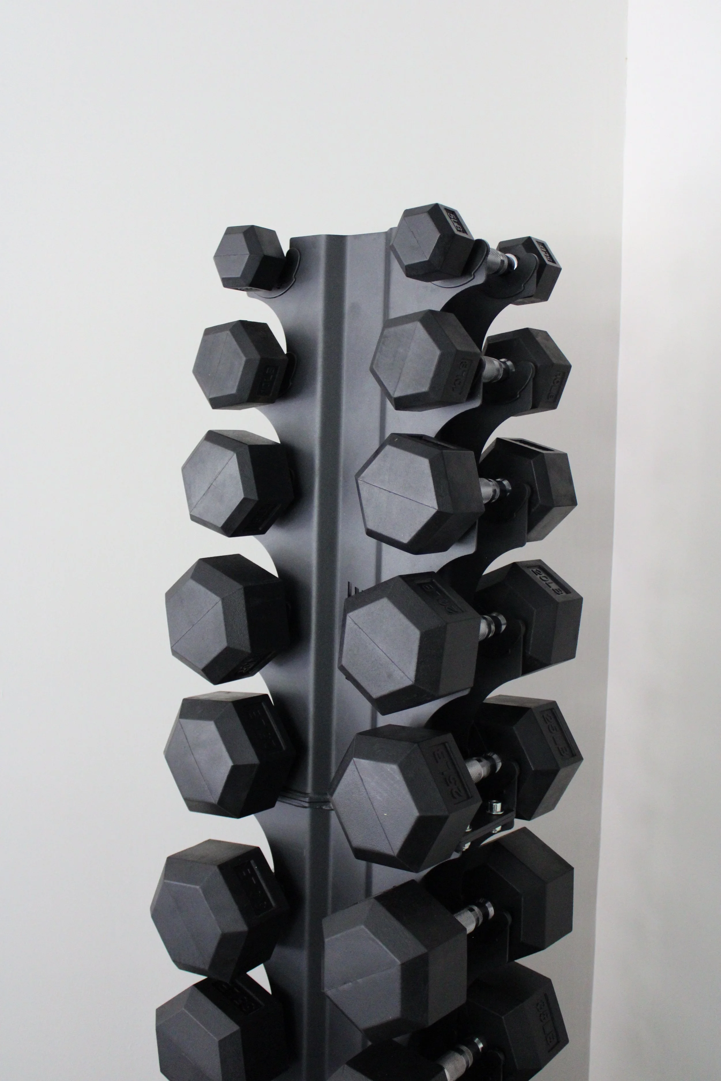 Standing rack of black and gray dumbbells on a gray vertical holder against a white wall.