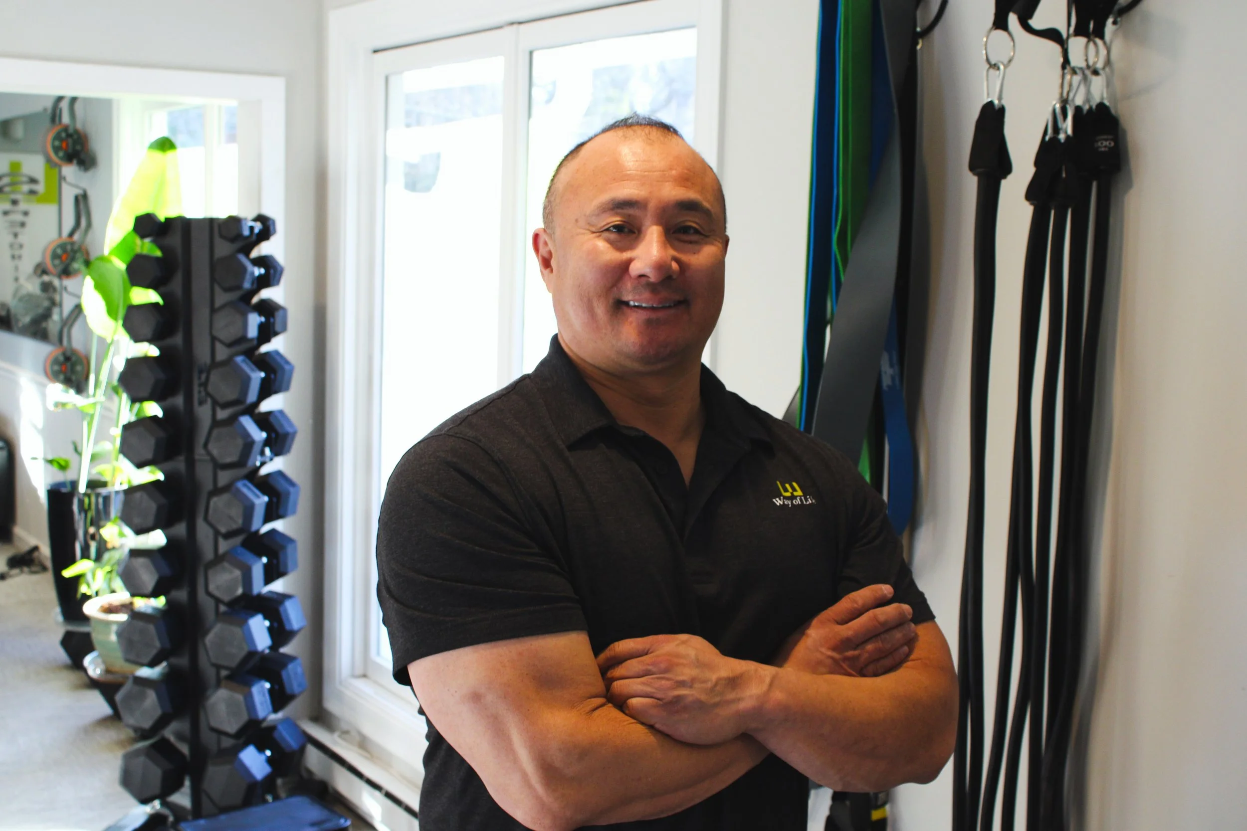 Man smiling with arms crossed in workout room with dumbbells, resistance bands, and a mirror.