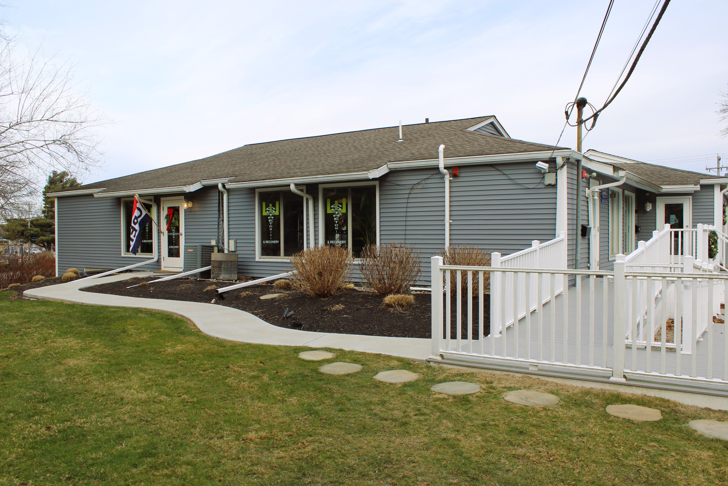 A gray commercial building with a ramp and white railing, decorated with small wreaths, and windows displaying 'Substance Recovery' signs, surrounded by landscaped grass and shrubs.