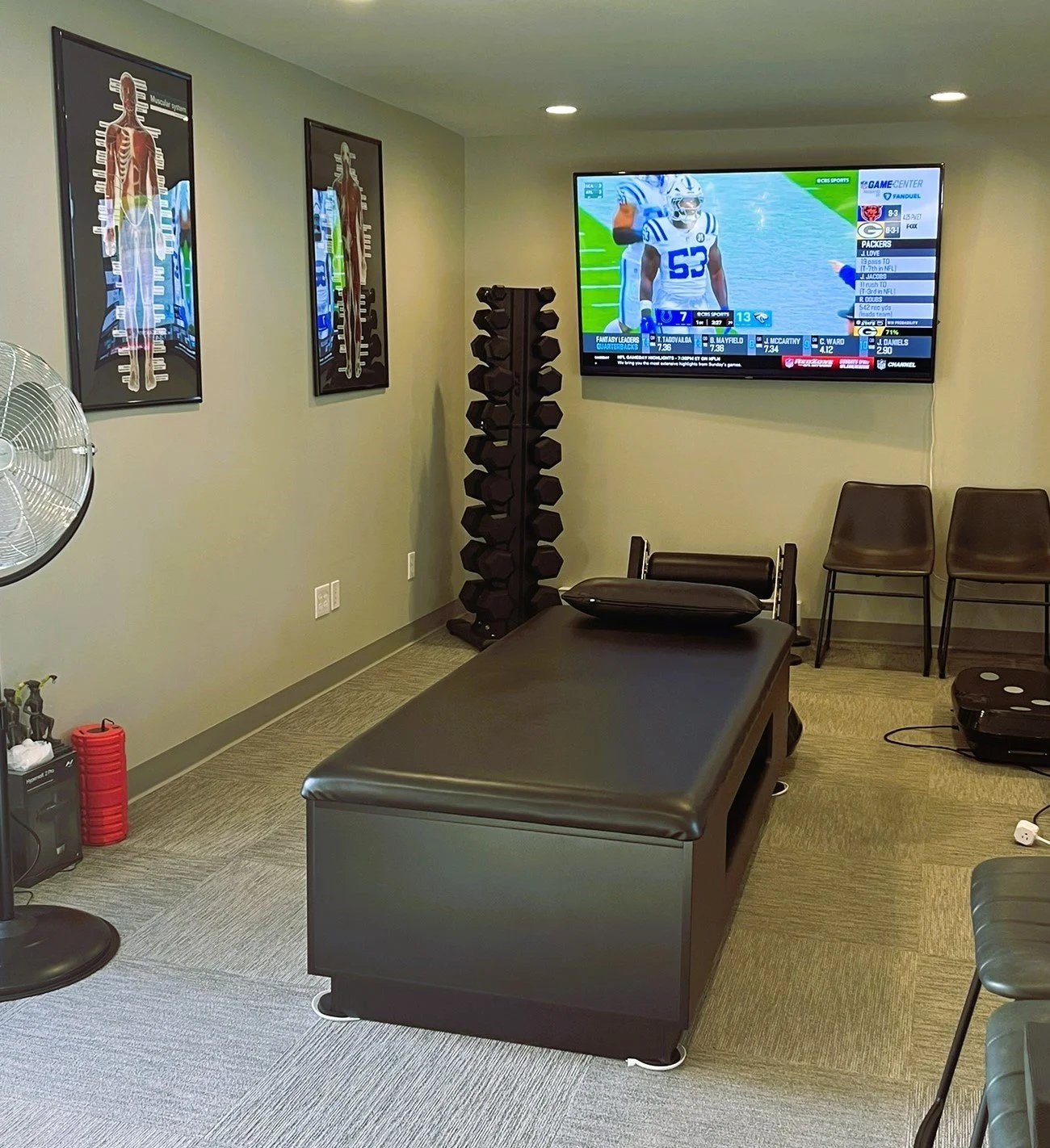 A chiropractor's examination room with a black padded table, a window fan, framed anatomy posters on the wall, a stack of black dumbbells in a rack, a large wall-mounted television displaying a football game, some chairs, and a black speaker on the floor.