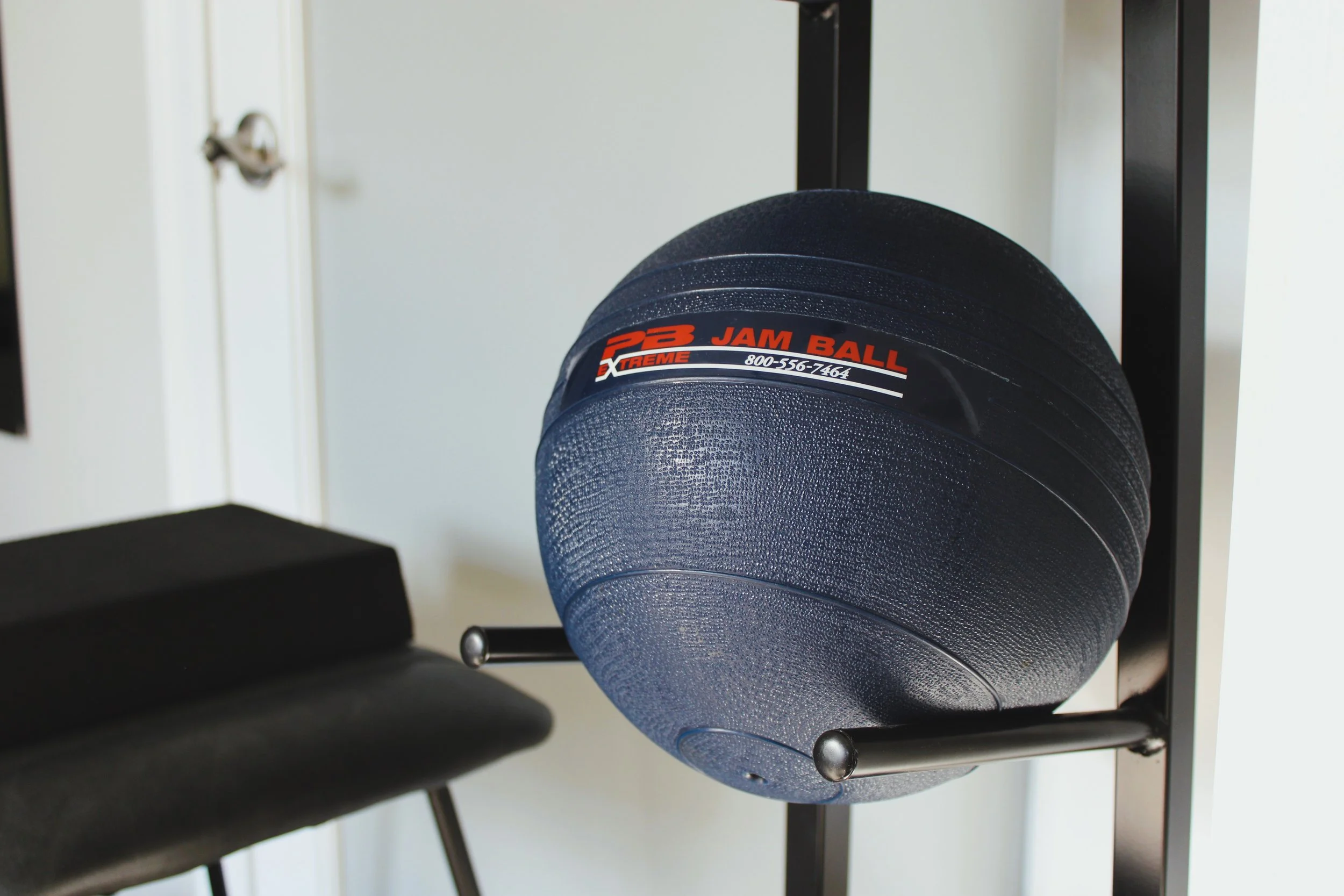 A black medicine ball labeled 'JAM BALL' with a phone number on a stand in a gym or workout space.