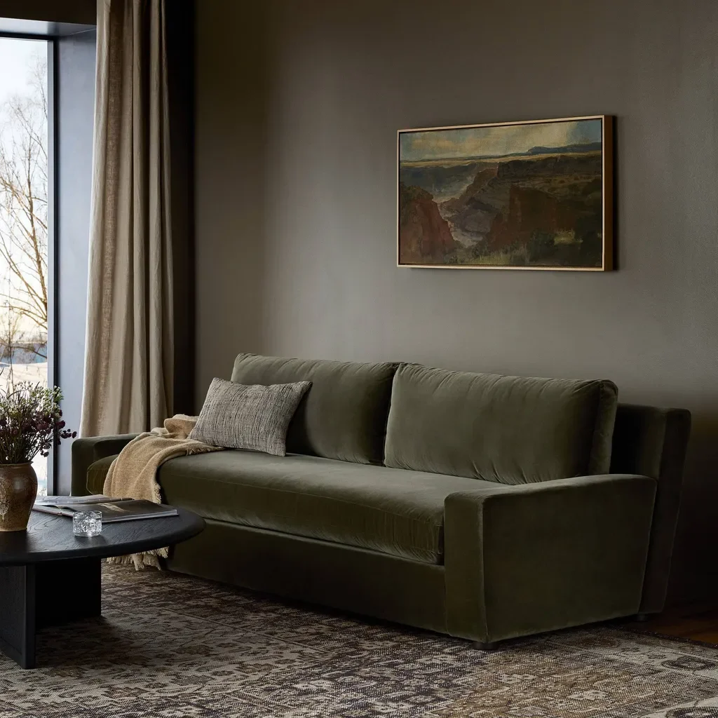 Living room with a green sofa, a framed landscape painting, a window with beige curtains, and a coffee table with a vase of flowers, a book, and a candle.