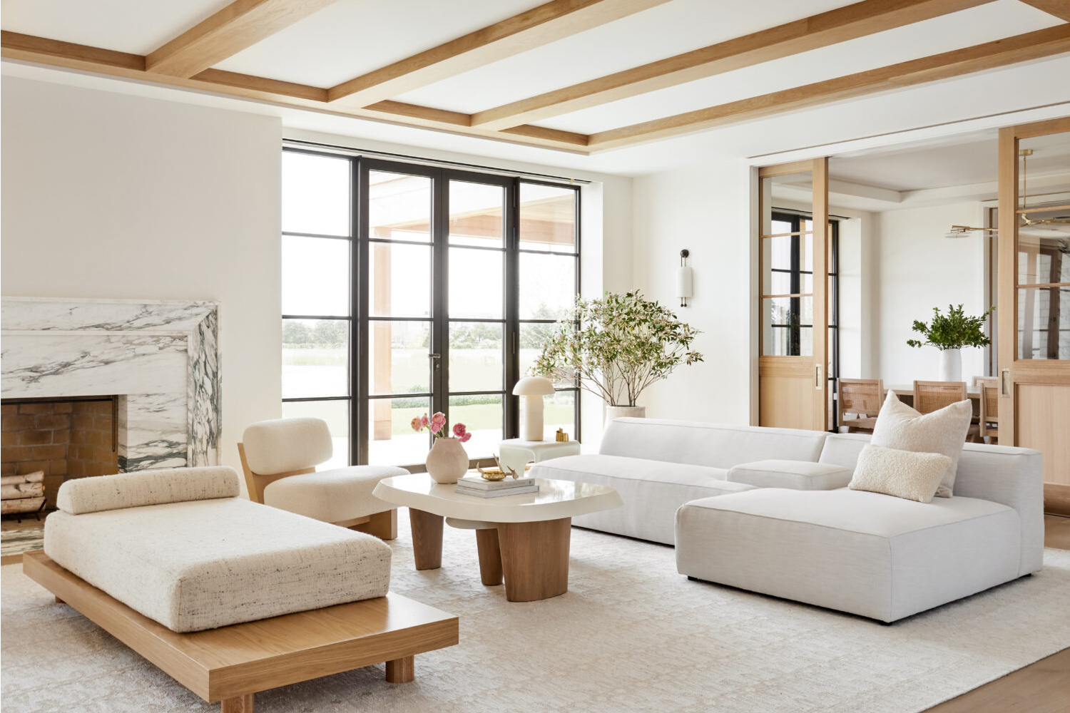 Modern living room with large windows, white sofas, a marble fireplace, wooden beams, and greenery.