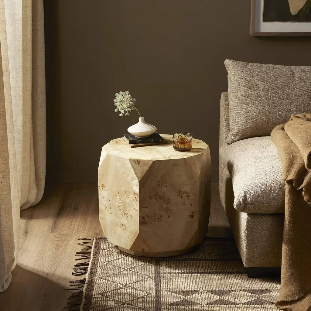 A beige, geode-style side table with decorative items on top, including a white vase with a small branch, a closed book, and a glass of whiskey, next to a gray upholstered armchair in a cozy living room.