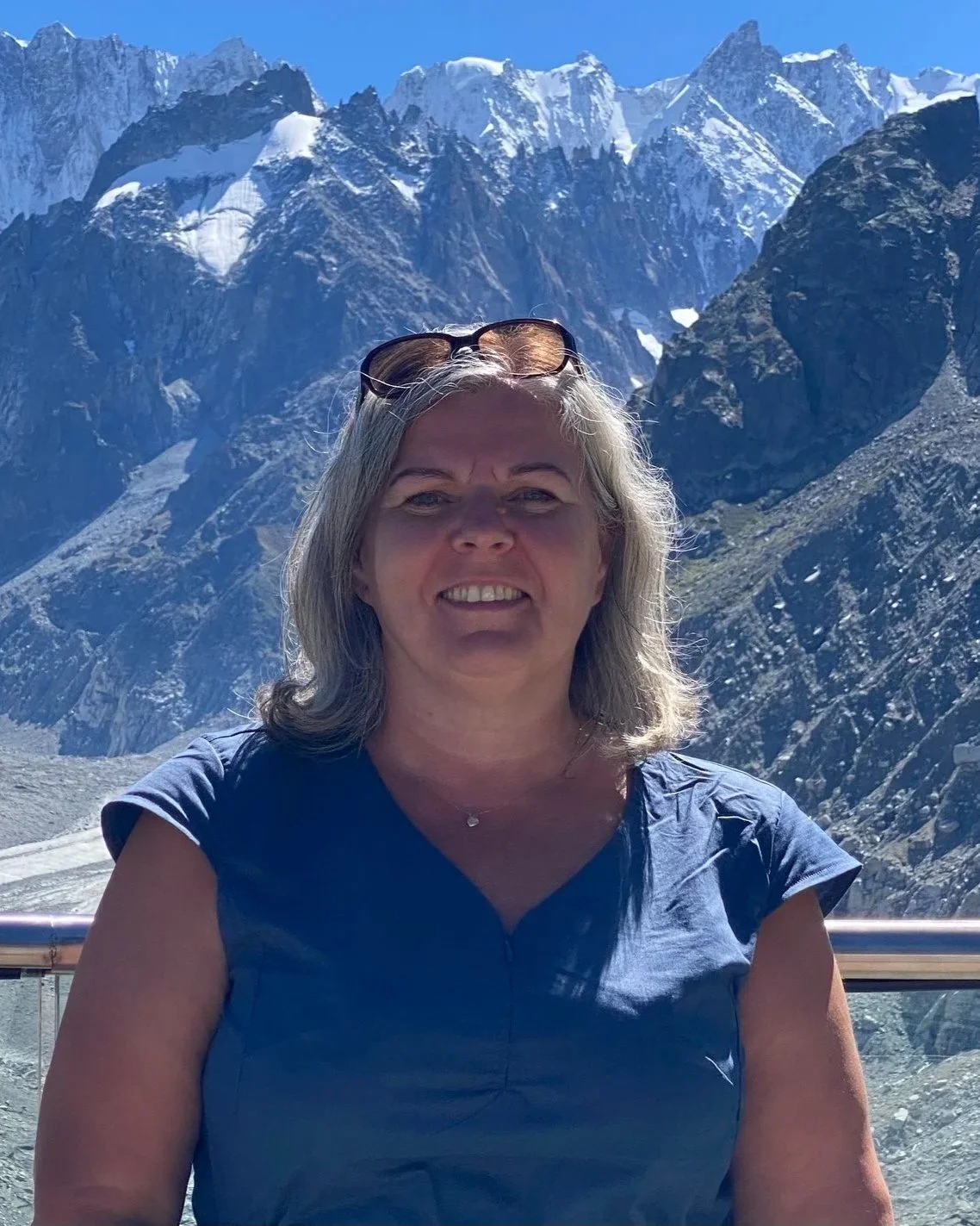 A woman with gray hair smiling, wearing a blue shirt and sunglasses on her head, standing in front of a mountainous landscape with snow-capped peaks under a clear blue sky.