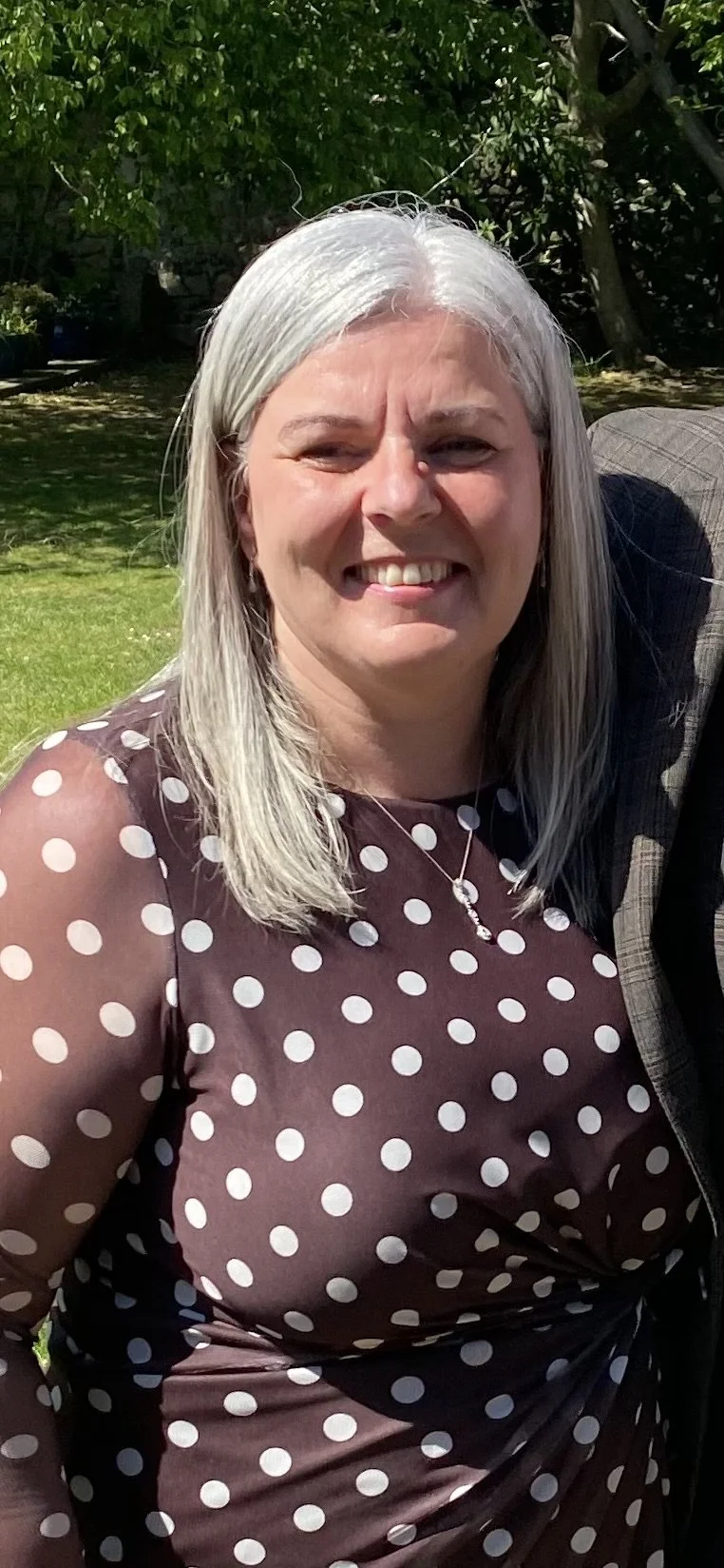 A woman with gray hair smiling outdoors on a sunny day, wearing a dark polka dot dress and a necklace, standing in a garden with green trees and grass.