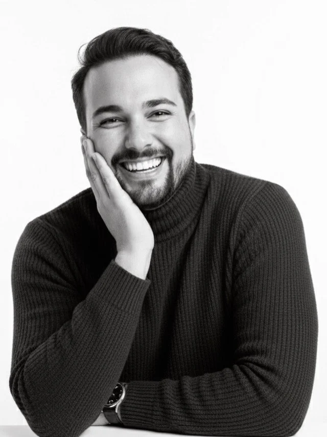 Black and white portrait of a smiling man with a beard, wearing a turtleneck sweater, resting his face on his hand.