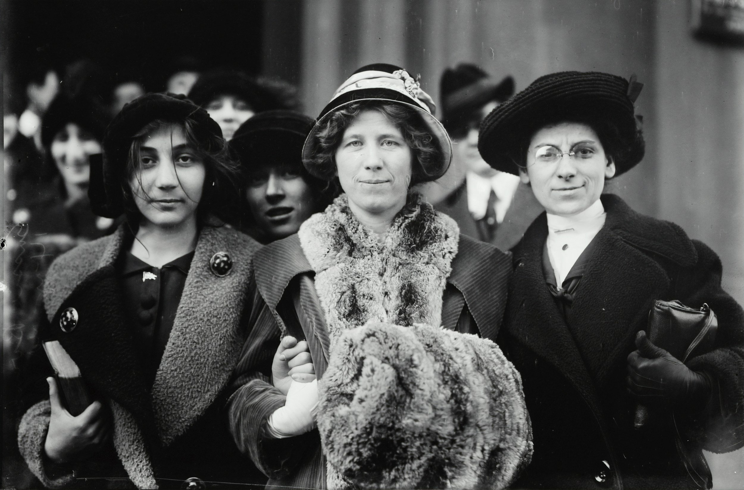 Black-and-white historical photograph of three women standing closely together in early 20th-century attire, wearing hats and heavy coats, with a crowd gathered behind them.