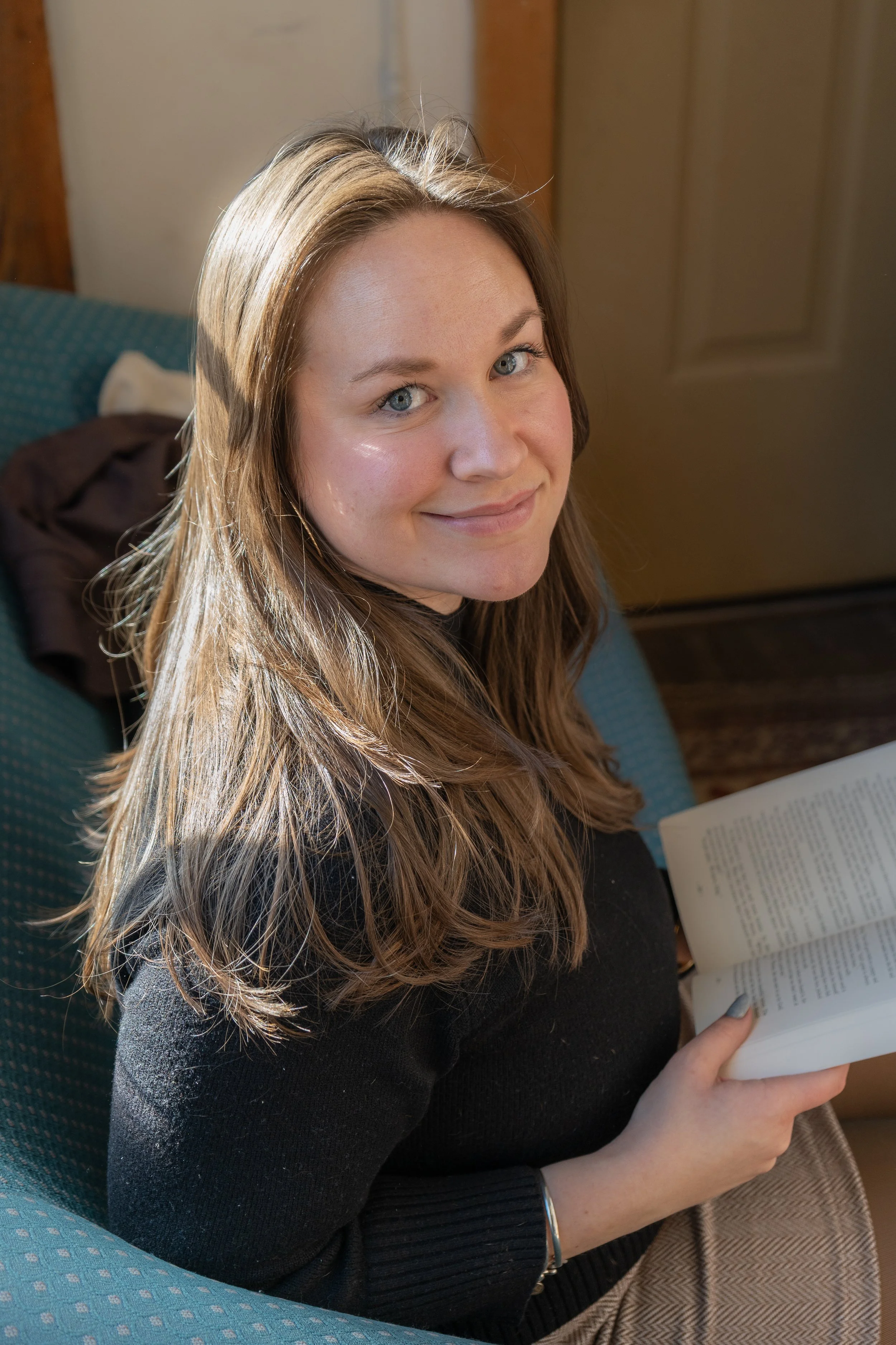 Young woman with long brown hair and blue eyes sitting on a patterned teal chair, holding an open book, smiling at the camera in a cozy indoor setting.