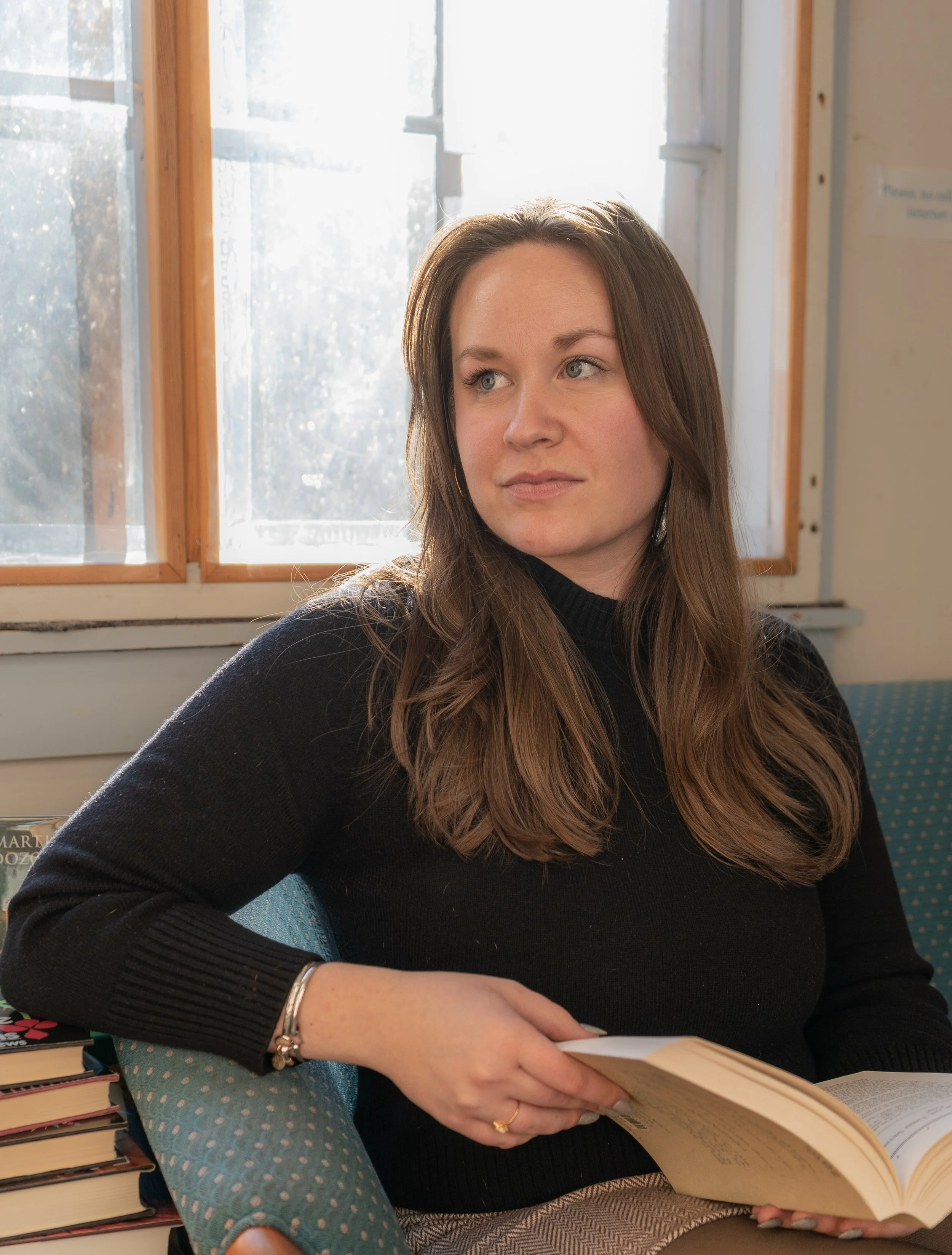A woman with long brown hair wearing a black turtleneck sweater sitting on a polka dot armchair holding an open book, with a stack of books next to her, in a room with sunlight coming through a window.