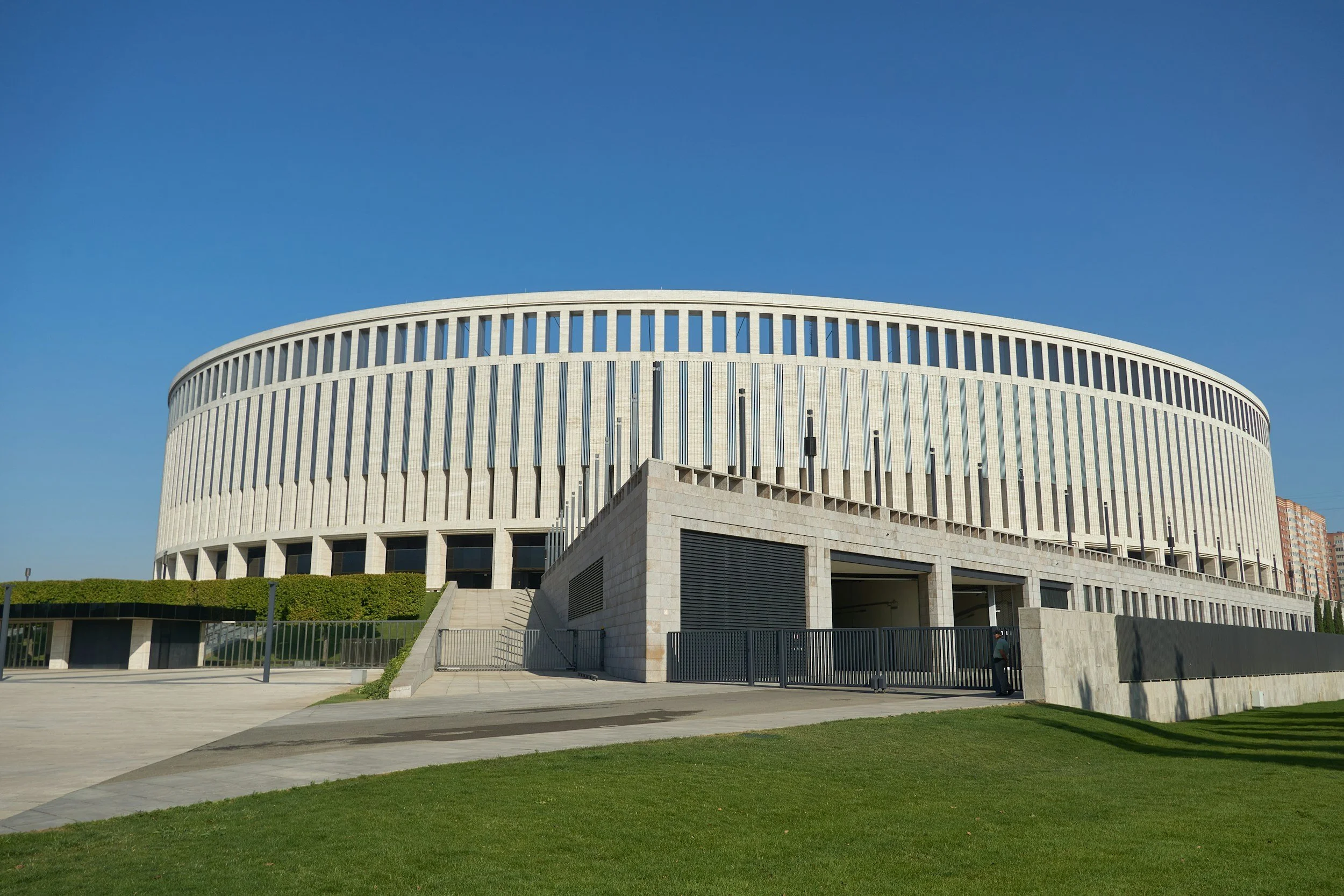 A large circular modern building with vertical white slats, a concrete staircase, green bushes, and a lawn under a clear blue sky.