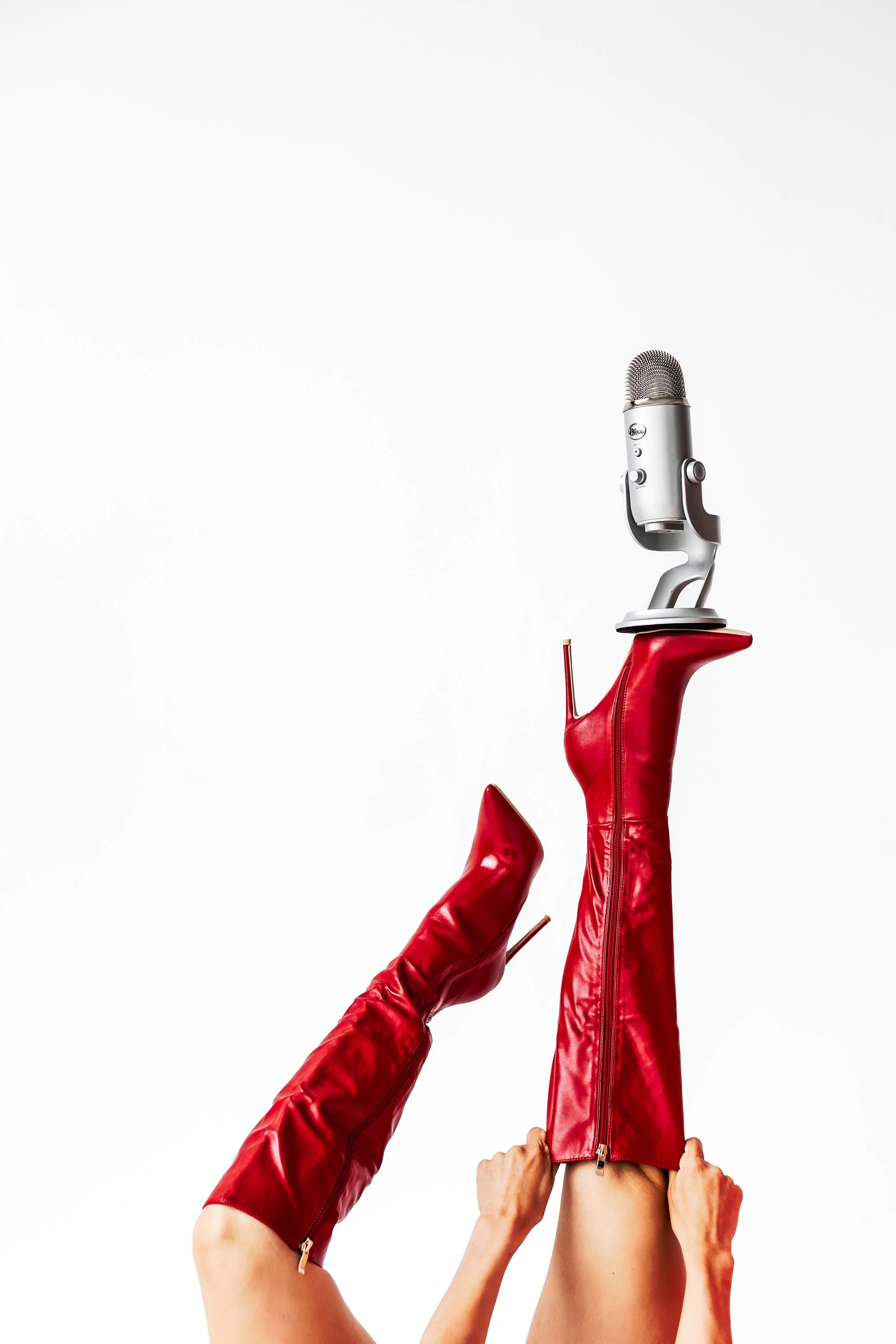 A person wearing red leather high-heeled boots and red leather pants or dress is holding a silver microphone above their head against a plain white background.