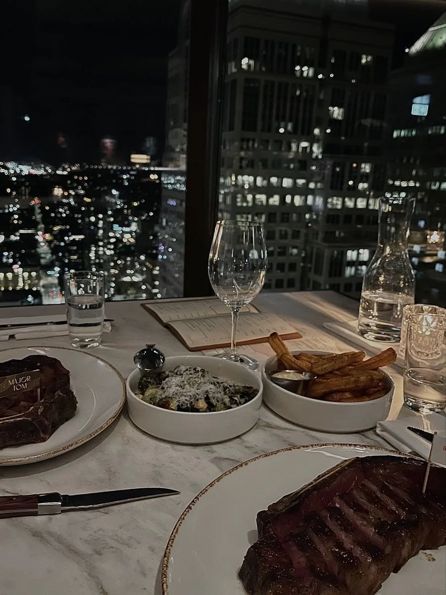 A dinner table at night with a city view, set with plates of steak, grilled fries, salads, water glasses, a wine glass, and a skillet, with menus and utensils.