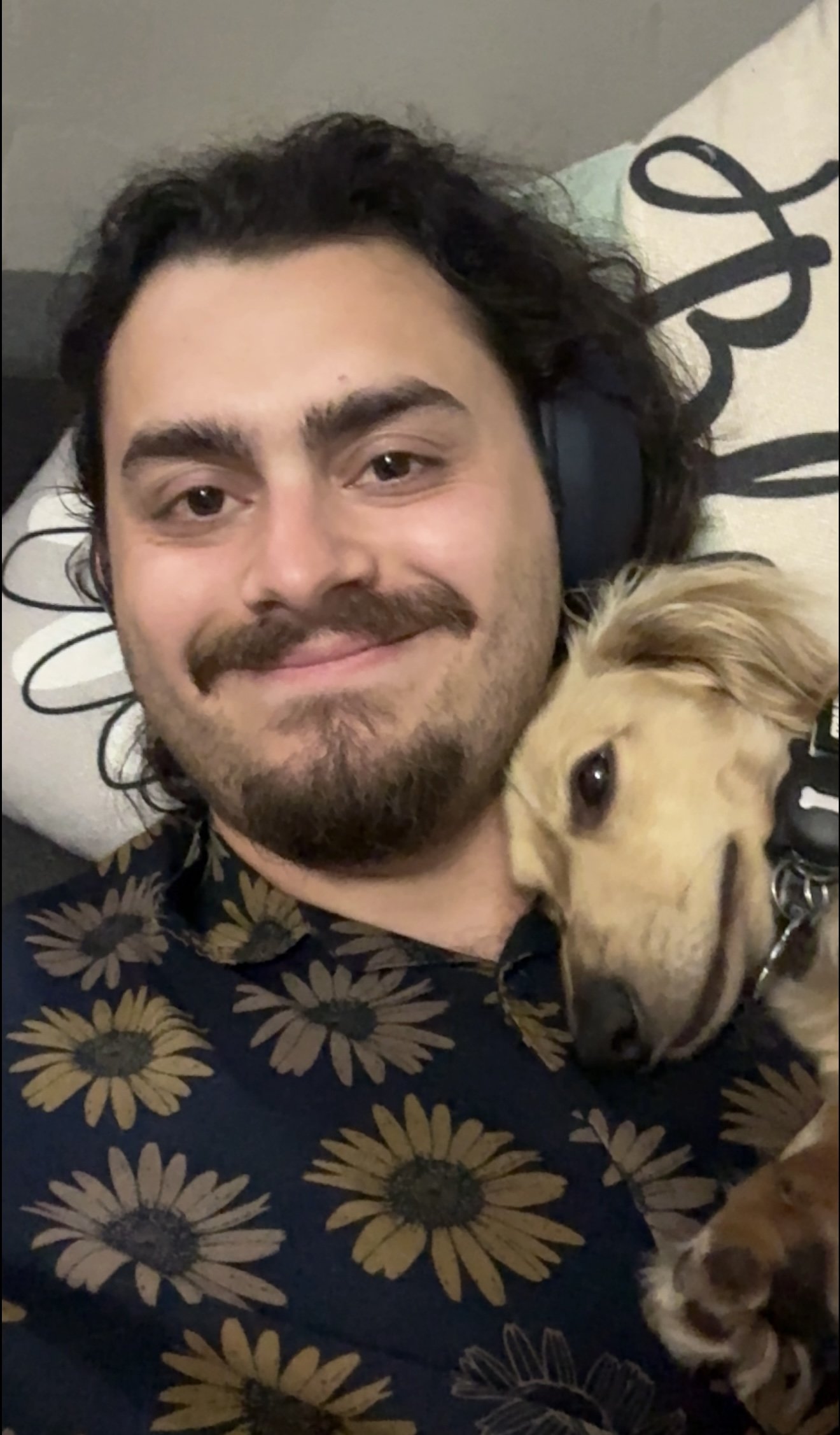 A man with dark wavy hair, a mustache, and a goatee, wearing a floral shirt, lying on a pillow with a black and white design, smiling, with a golden retriever puppy resting its head on his chest.
