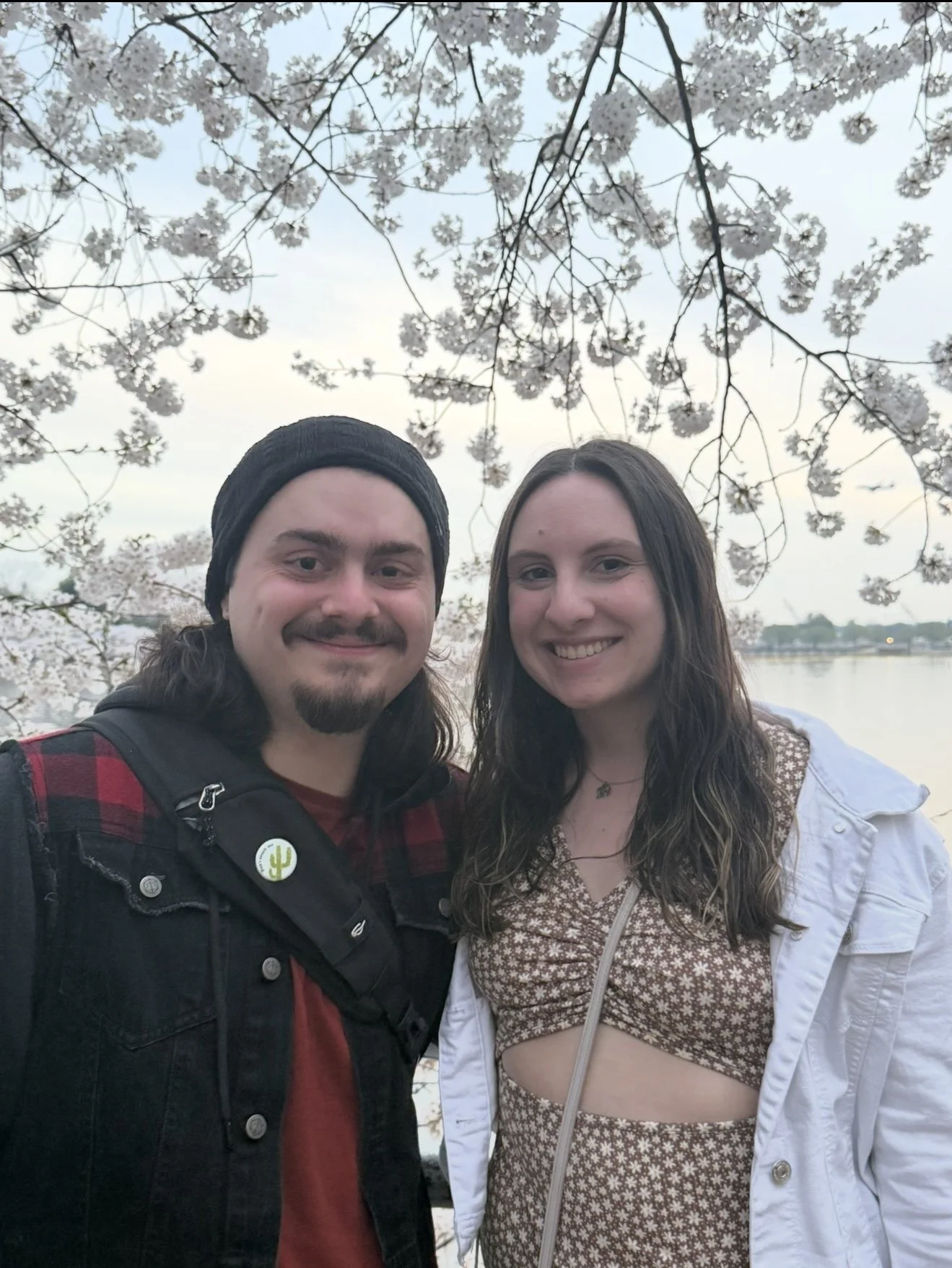 A young man and woman taking a selfie outdoors near a body of water, with cherry blossom trees in bloom overhead.