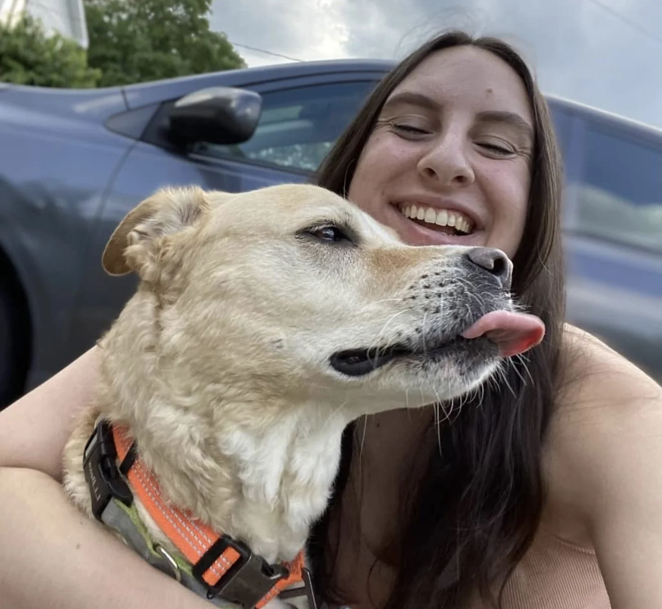 A woman smiling while taking a selfie with her dog, which has its tongue out, in front of a parked car.