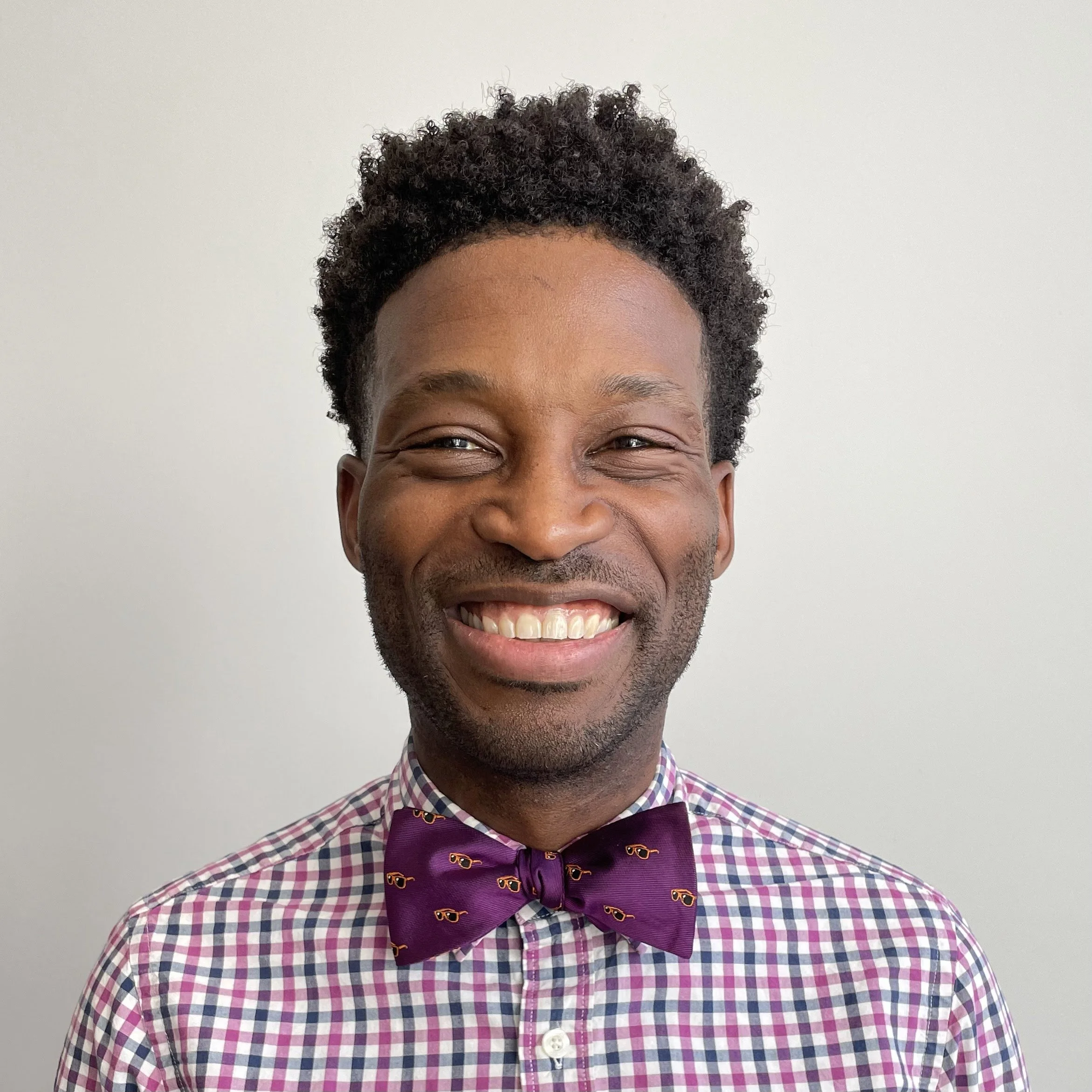 A smiling man wearing a purple bow tie with sunglasses pattern, a checkered shirt, standing against a plain light background.