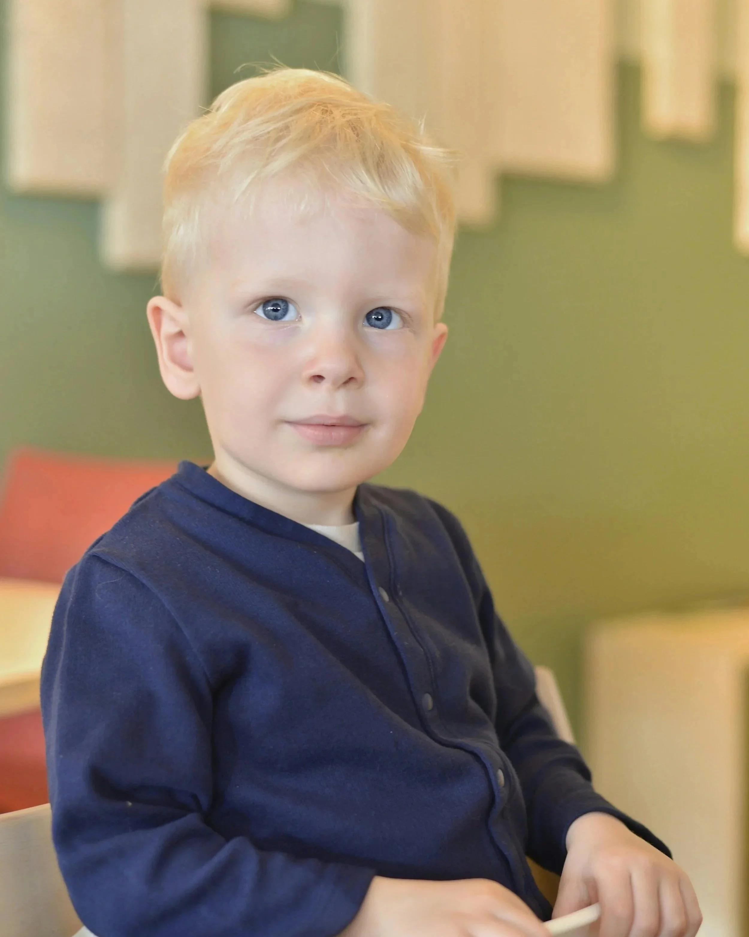 A young boy with blond hair and blue eyes sitting indoors, wearing a navy blue long-sleeve shirt, looking at the camera.
