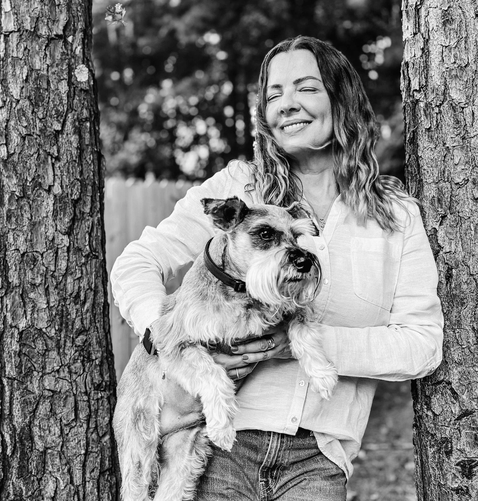 A woman smiling with eyes closed holds a dog between two tree trunks outdoors.