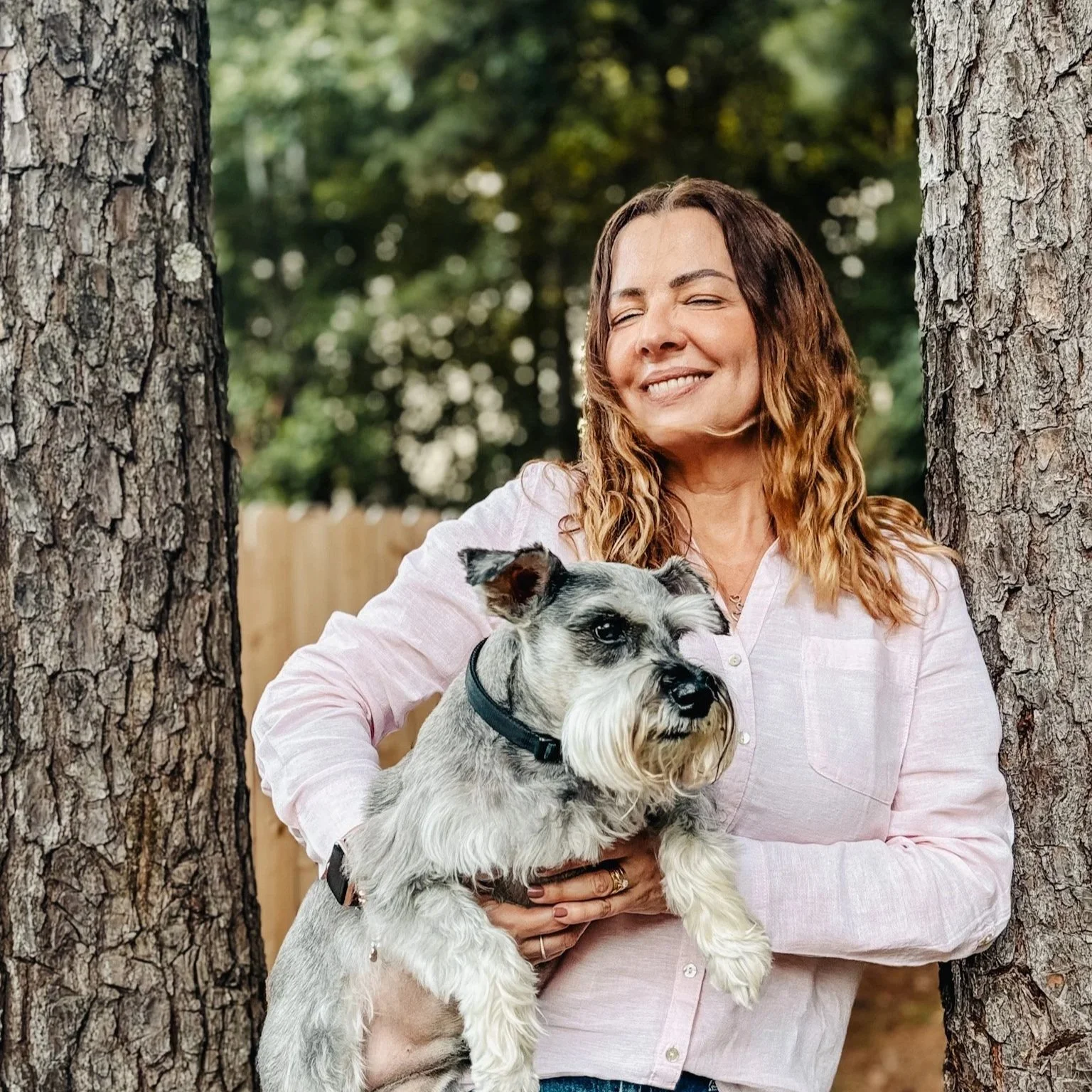A woman with wavy brown hair smiling with eyes closed, holding a gray and white Schnauzer dog between two trees in a backyard.