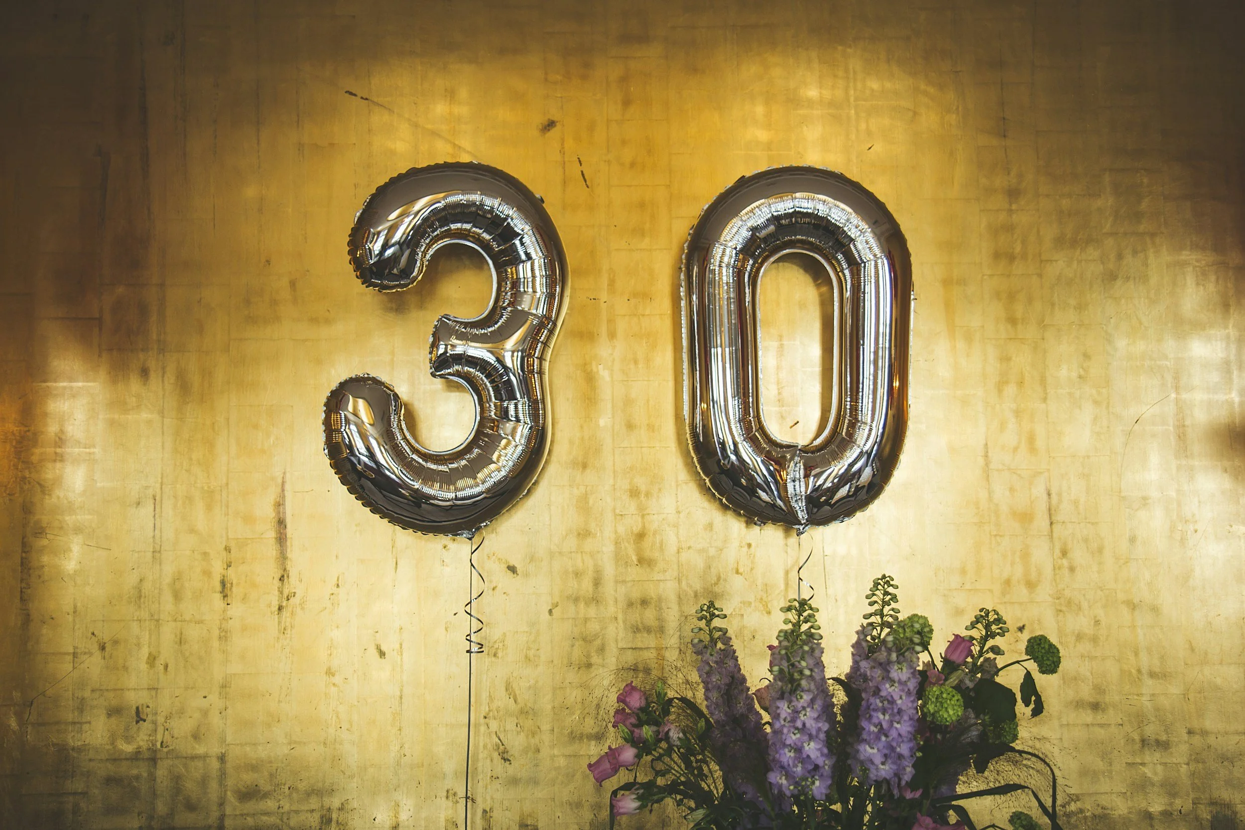 Silver number balloons forming '30' on a gold wall with a bouquet of purple and green flowers below.