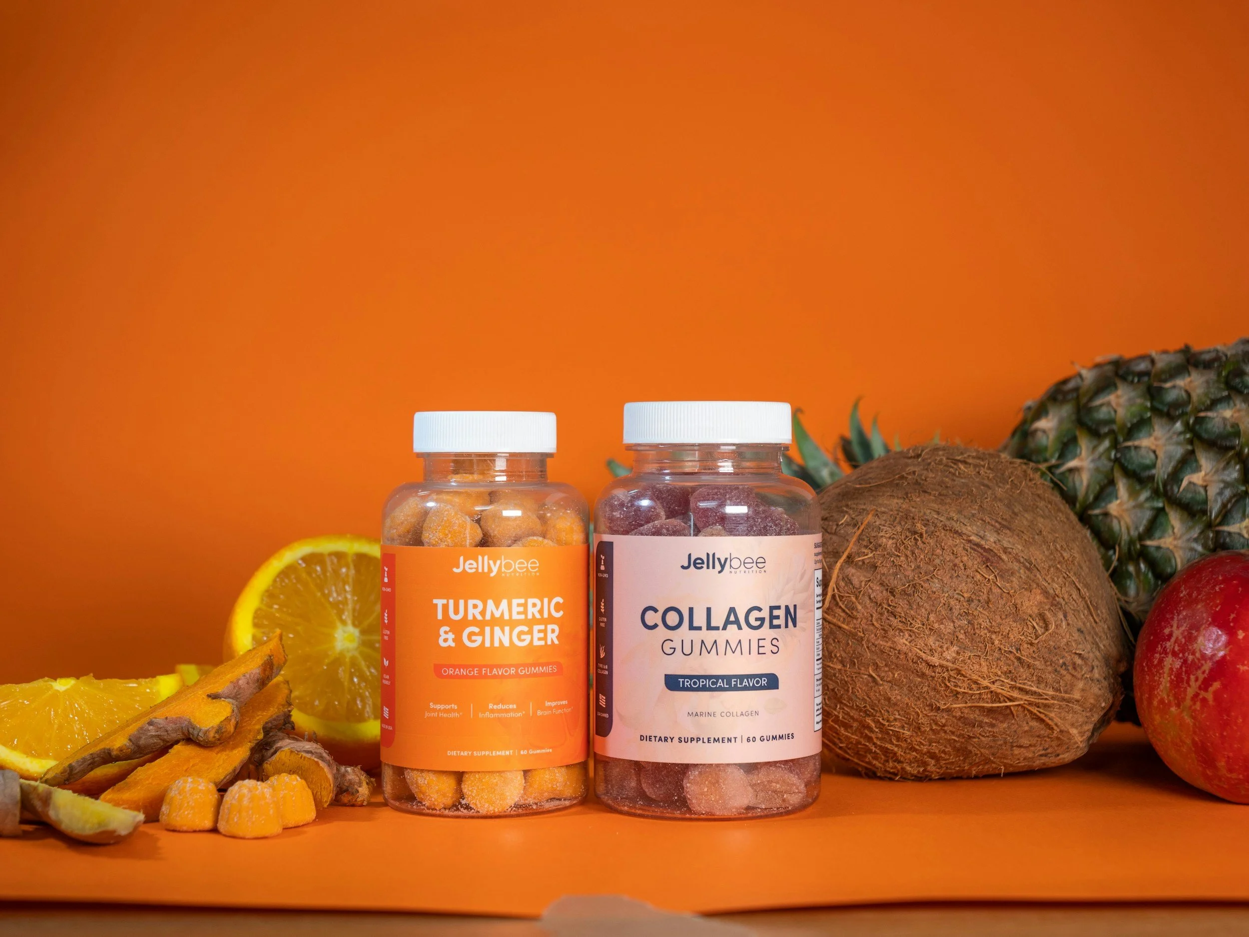 Two bottles of dietary supplements, one labeled 'Turmeric & Ginger' and the other 'Collagen Gummies,' placed among sliced orange, turmeric root, coconut, pineapple, and apple on an orange background.