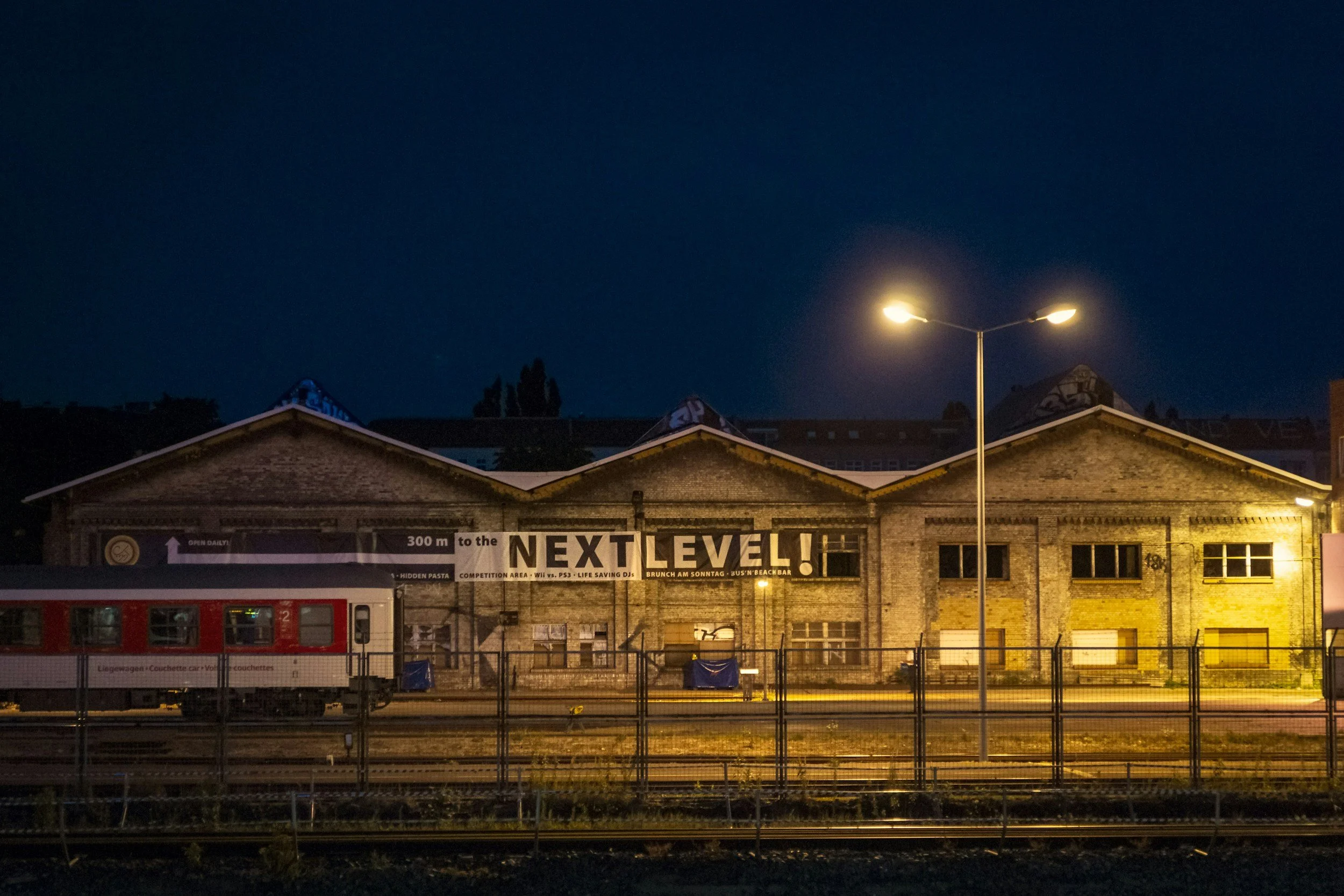 A nighttime scene shows the roof of a brick building with signage that reads 'Next Level!' and other advertising text. There is a streetlamp illuminating the area and a red train car on tracks in the foreground. The sky is dark with some stars visible.
