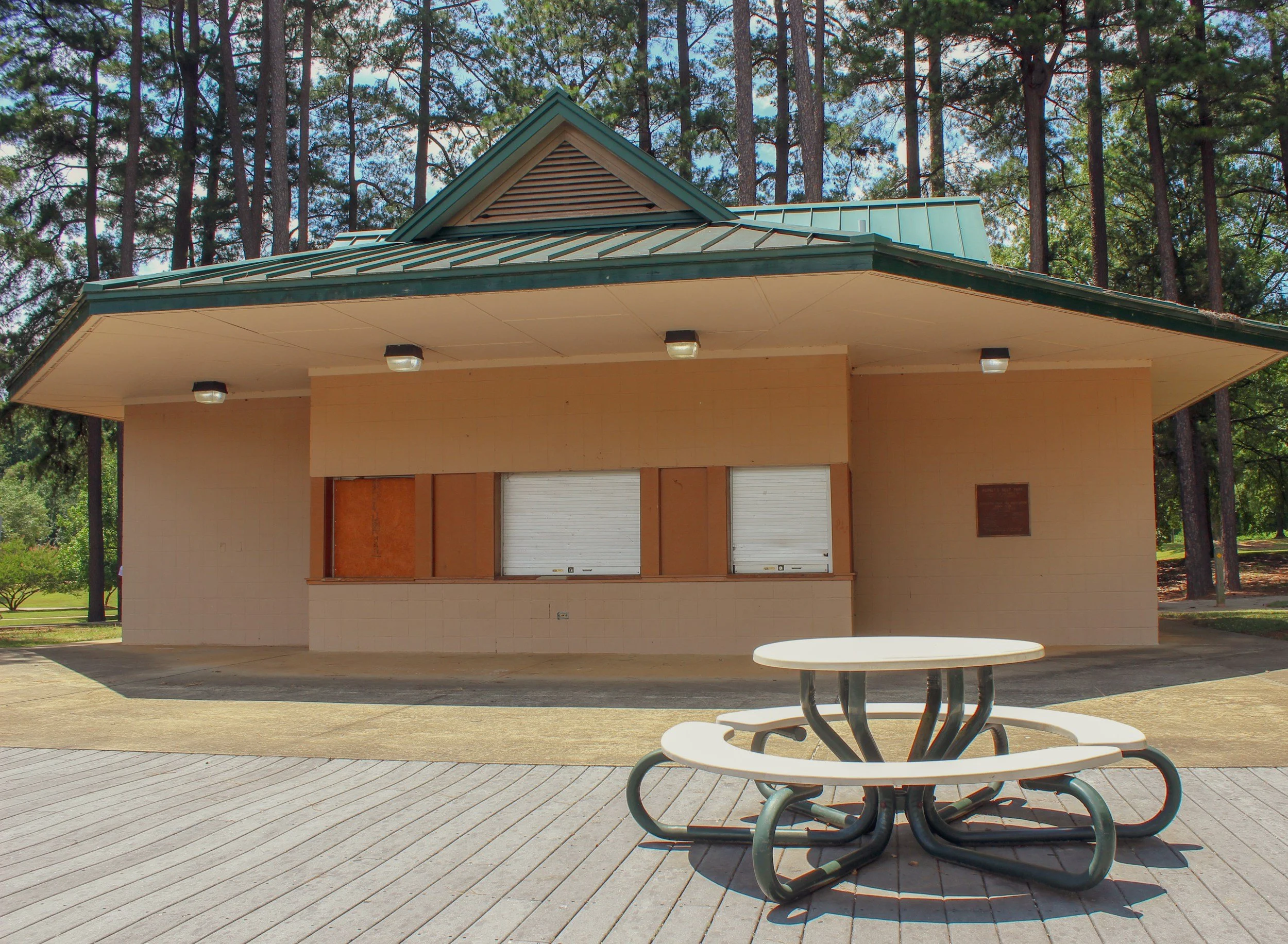 A wooden building with a green metal roof and shutters, surrounded by tall trees, with an outdoor table and benches in the foreground.