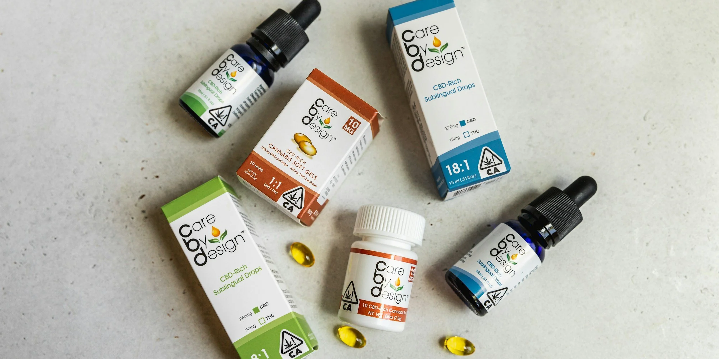 Assorted CBD and cannabis product packaging, including dropper bottles, soft gels, and sublingual drops by Care by Design, on a light-colored surface.