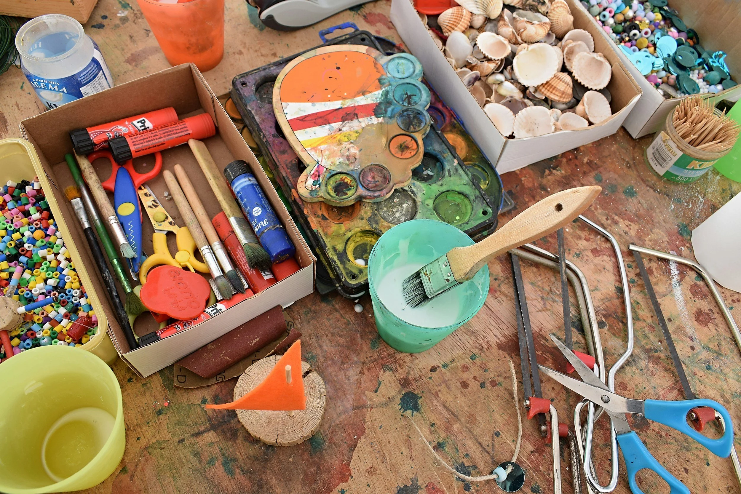 A cluttered table with arts and crafts supplies, including shell collections, paint palette, brushes, glue sticks, scissors, pipe cleaners, and foam cups.