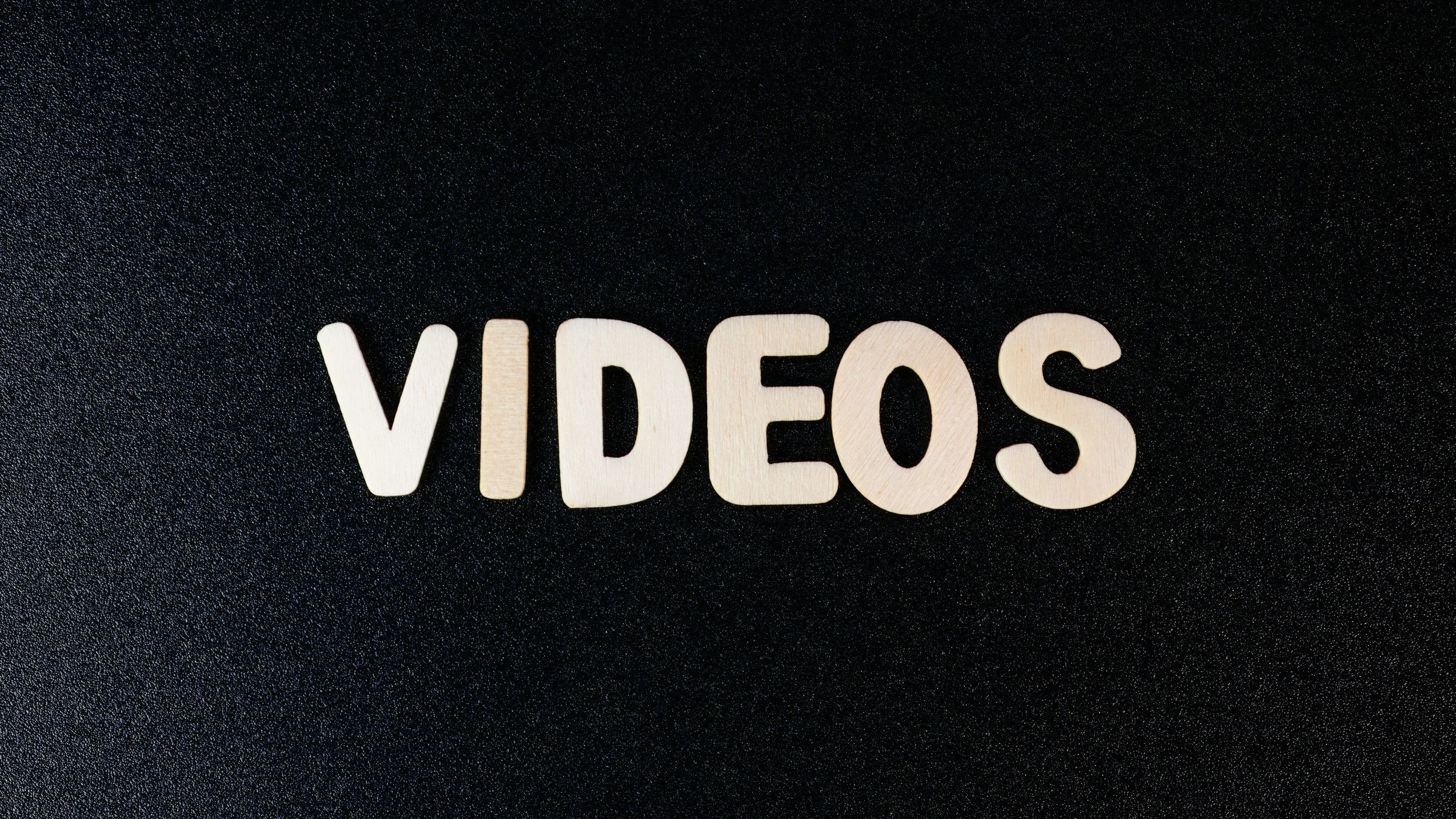 The word 'VIDEOS' spelled out with light-colored wooden letters on a black textured background.