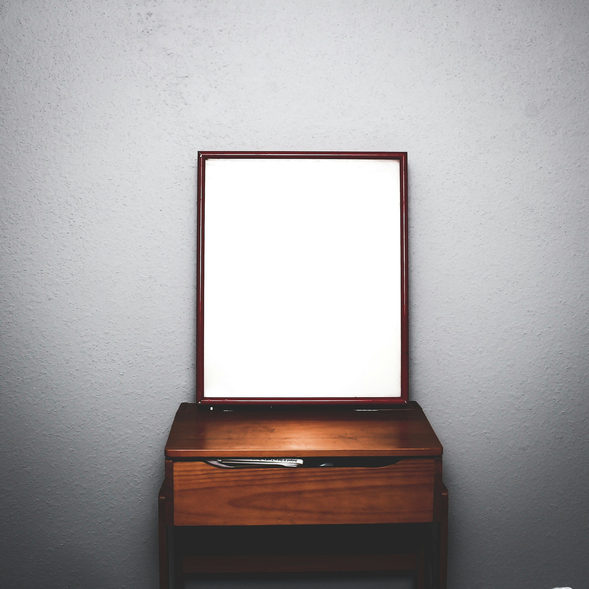 A wooden table with a rectangular mirror with a red frame leaning against a white textured wall, illuminated by a warm light.