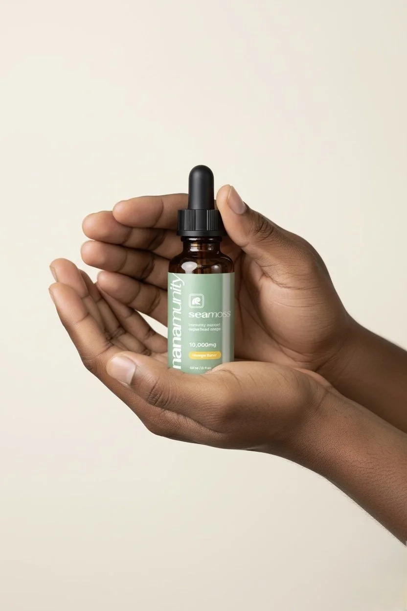 A person with dark skin holds a small amber glass bottle with a black dropper cap. The bottle has a green label with the word 'mananality' and descriptions indicating it contains 10,000 mg of a product called 'Seamoss,' which is described as an immunity and energy-boosting supplement.