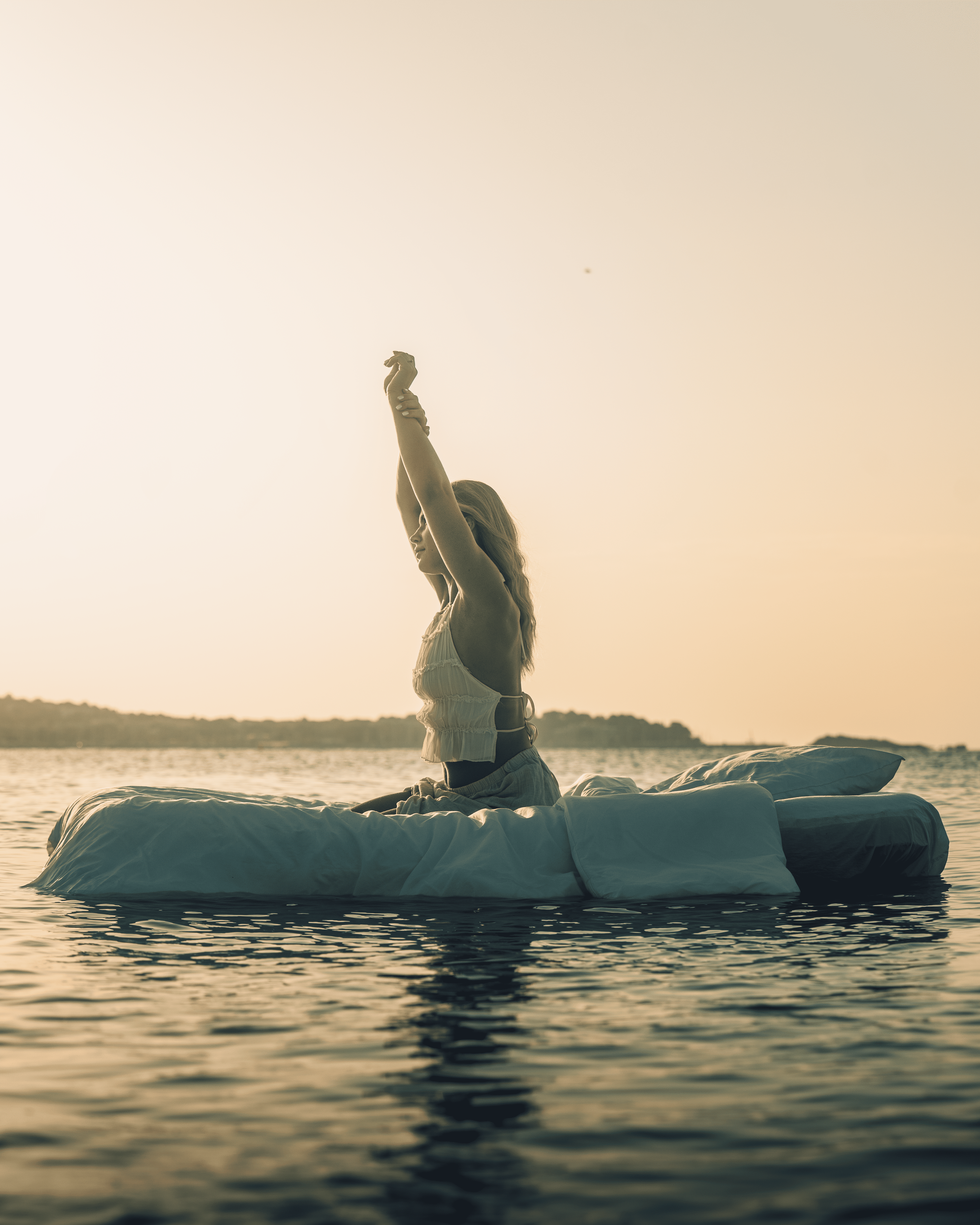 Femme assise sur un matelas flottant dans l'eau, faisant un étirement au coucher du soleil.