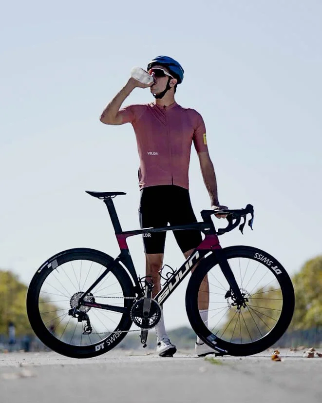 Cycliste en tenue de course, portant un casque bleu, buvant une bouteille d'eau avec un vélo de route noir et rose à ses côtés, sur une route en plein air.
