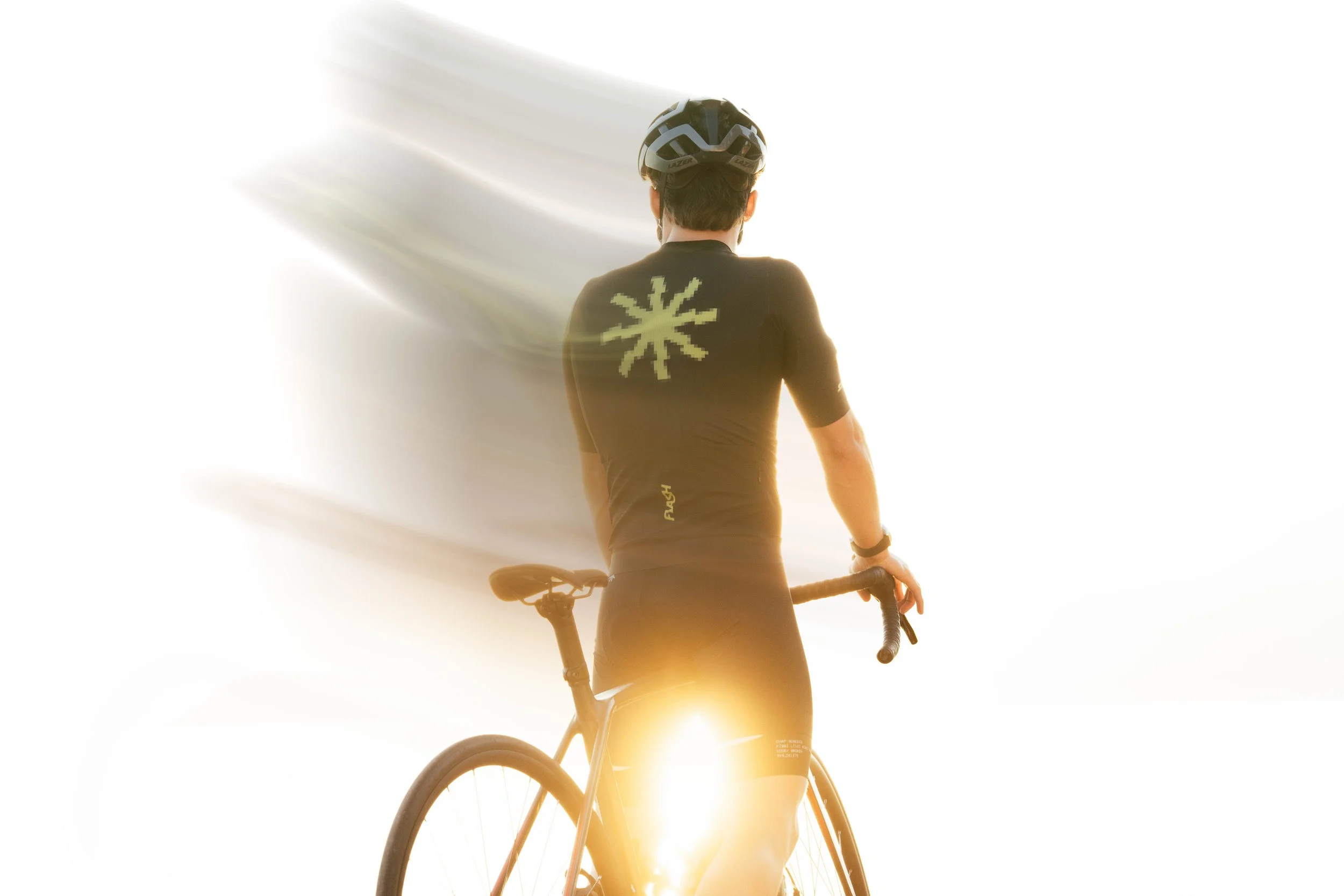 Un cycliste vu de dos, portant un casque et un maillot noir avec un motif en forme d'étoile sur le dos, tenant le guidon de son vélo avec l'éclat du soleil en arrière-plan.