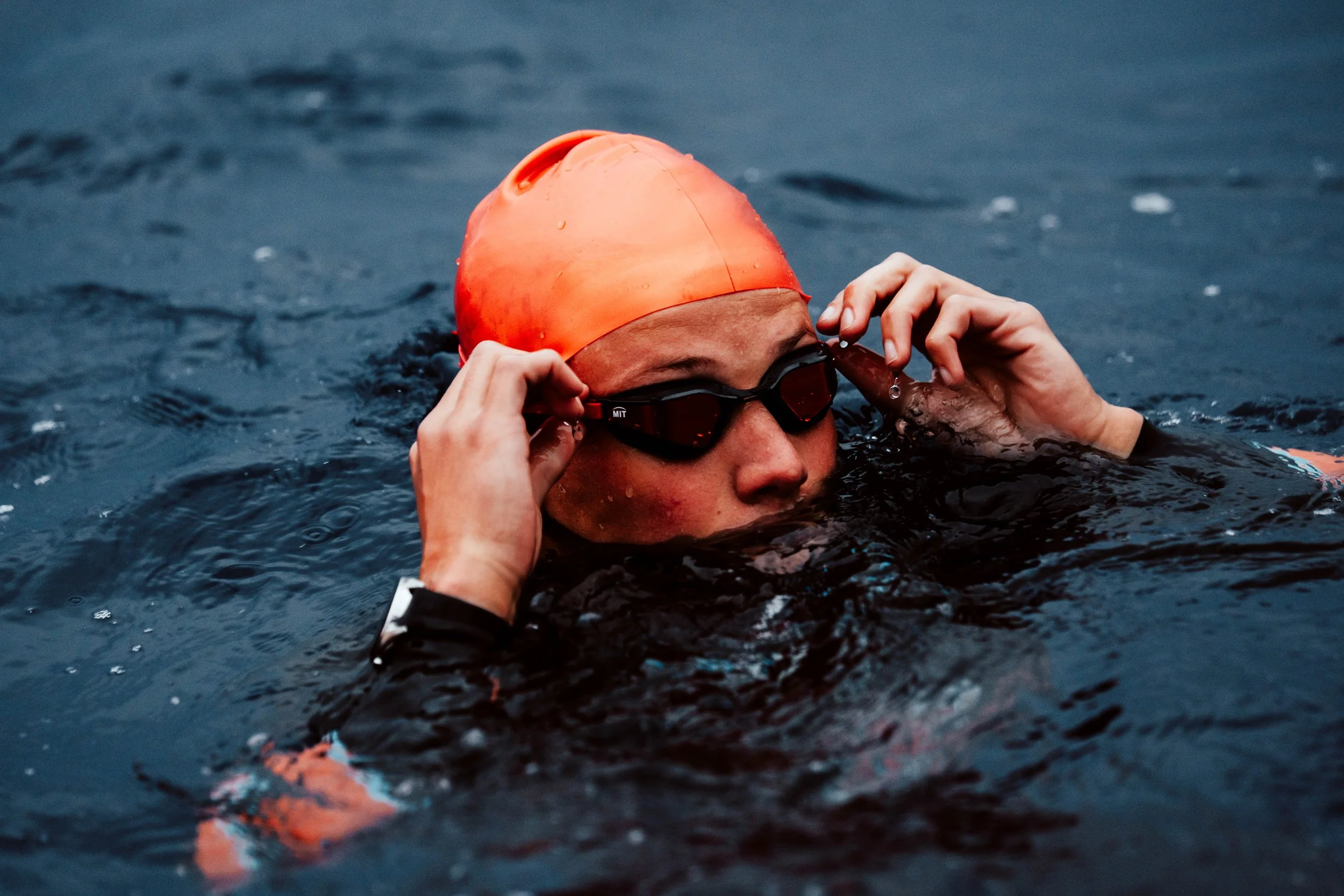 Nageur portant un bonnet orange et des lunettes de natation, sortant de l'eau dans une piscine ou un plan d'eau.