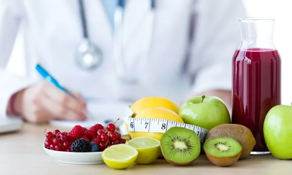 A healthcare professional's hand with a blue pen, a measuring tape, and a variety of fresh fruits including apples, kiwis, lemons, and berries on a wooden surface, with a bottle of juice. Nutrition