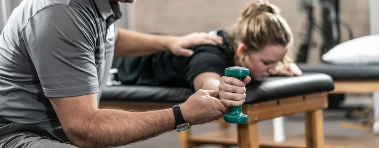 A woman lying face down on a massage table receiving physical therapy or chiropractic treatment, while a physical therapist uses a dumbbell to assist with a stretching or strengthening exercise. Rooted Spine & Sport Therapeutic Exercises