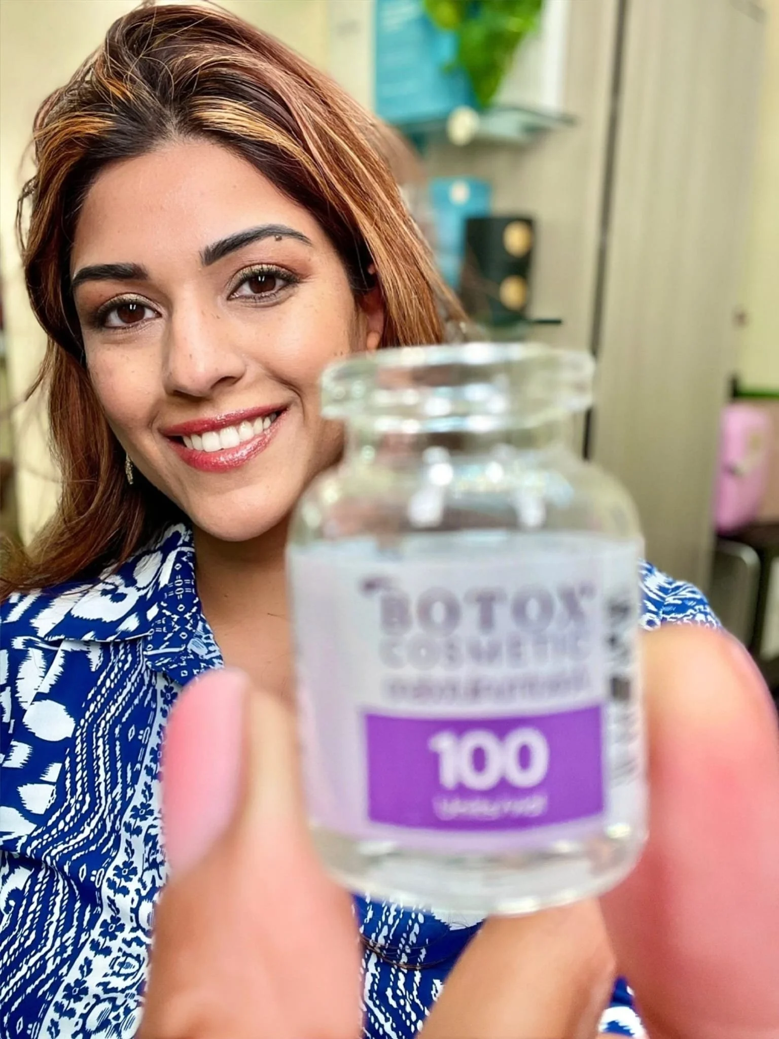 A smiling woman with brown hair and a blue patterned shirt holding a clear container labeled 'BOTOX COMPLEX 100 capsules' in front of her face in an indoor setting.