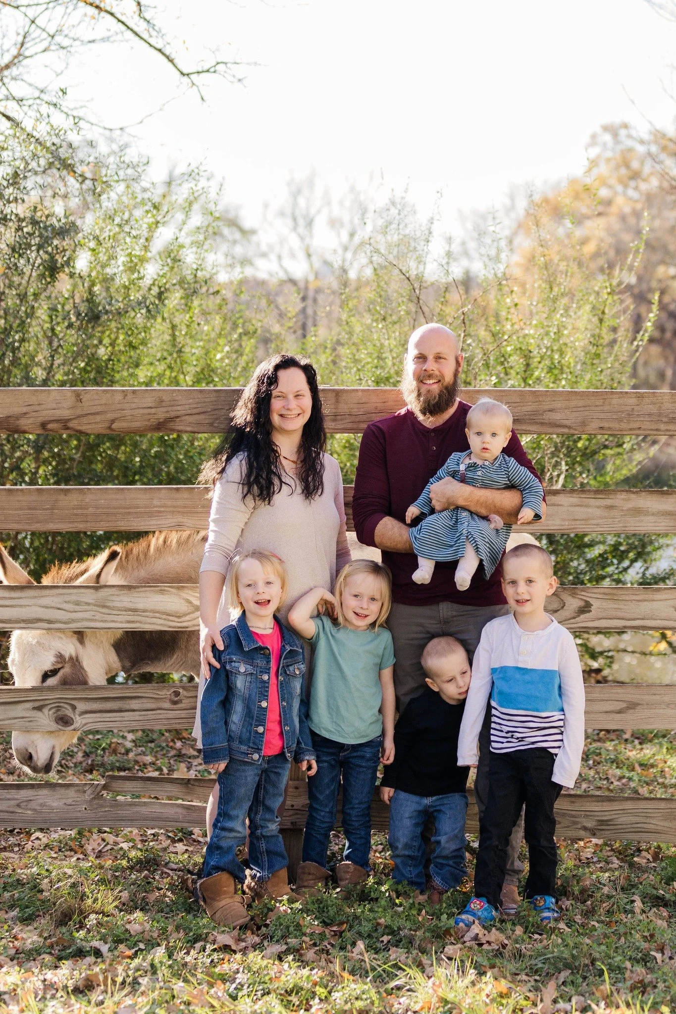 A family of seven stands in front of a wooden fence with a donkey behind it. The family includes two adults and five children, all smiling and posing outdoors on a sunny day with trees in the background.
