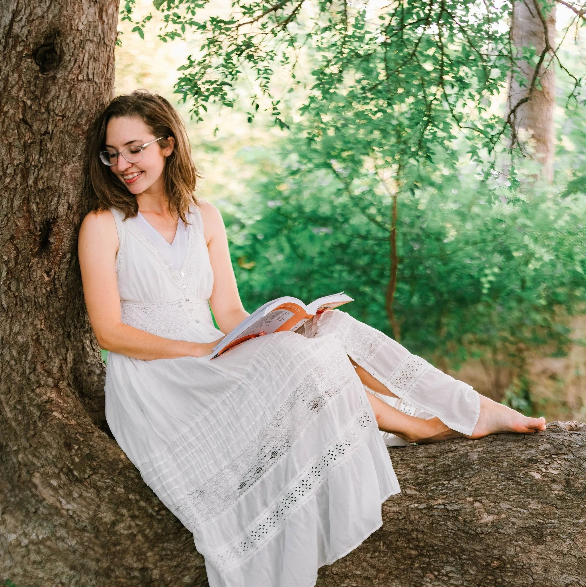 A young woman in a white dress with glasses is sitting on a tree branch, reading a book, in a green, leafy outdoor setting.