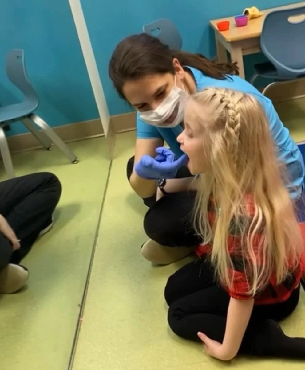 A young boy with brown hair wearing a salmon-colored t-shirt stands at a white table in a playroom, holding a small wooden box filled with sand and seashells. There are toys and decorative items on the table and in the background, including a stuffed turtle on the table, woven baskets, and a framed wall art piece.